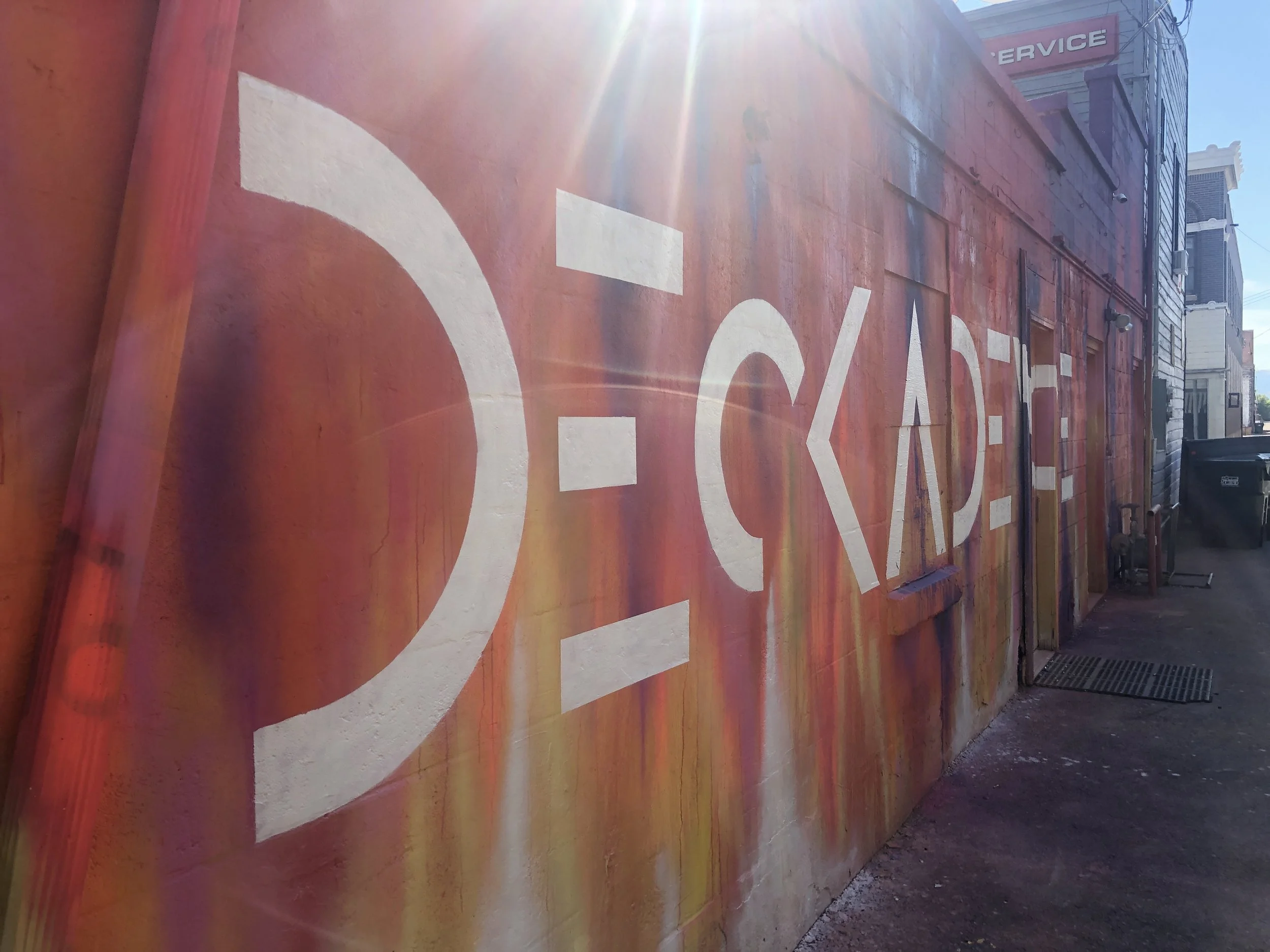 A red wall with white painted letters and a partial view of a street with buildings, electrical wires. The sunlight creates glare in the image.