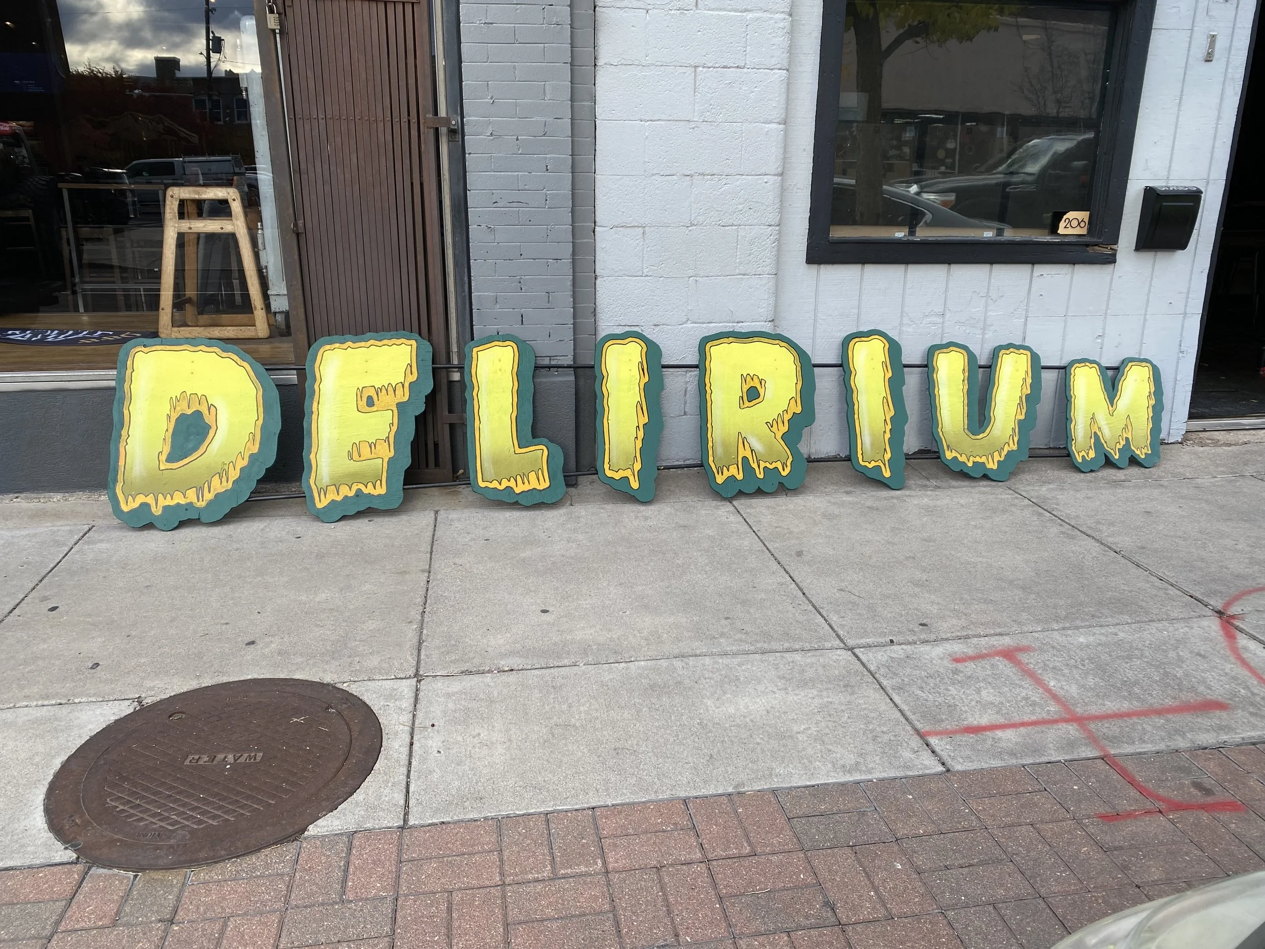 A sidewalk scene with large yellow block letters spelling 'DELIRIUM' placed in front of a building. The building has a window and a door, with a wooden stool visible through the window inside.