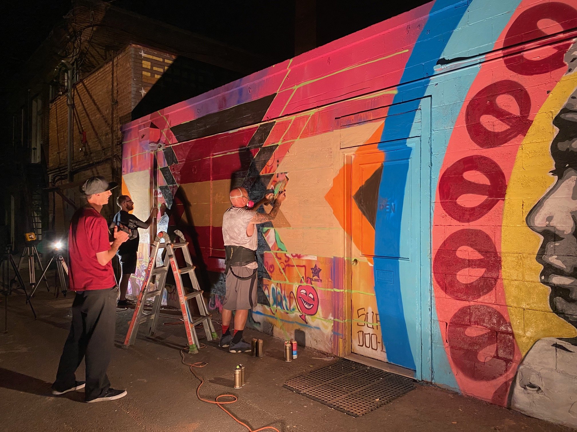 People painting a colorful mural on a large exterior wall at night. The mural features bold, bright colors, playful designs, and text.