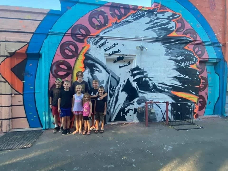 A group of seven children standing in front of a colorful mural of a Native American chief wearing a feather headdress on a city wall.