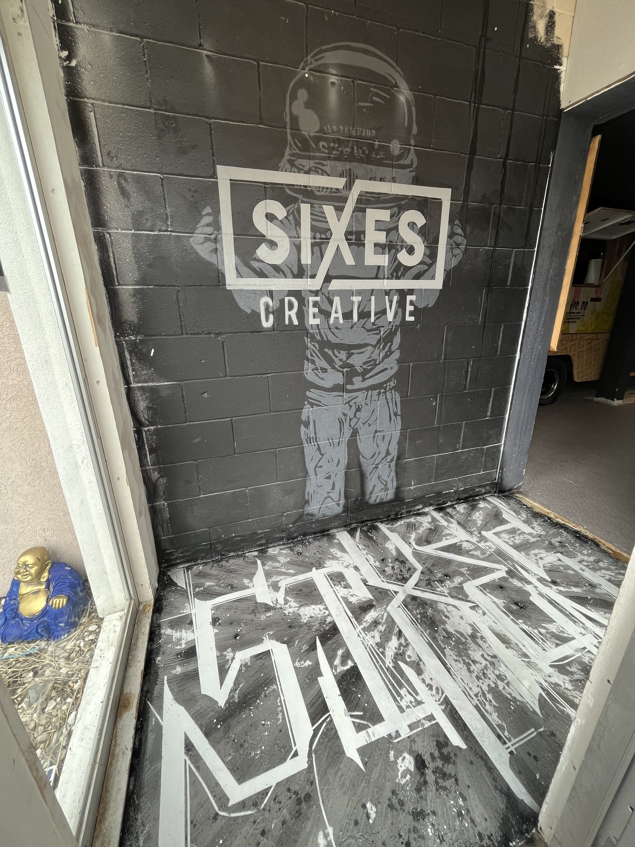 A black brick wall with a logo that says 'SIXES CREATE' and an illustration of a person in a hoodie with their face covered, standing with crossed arms. The wall and logo are located at the entrance of a store or business.