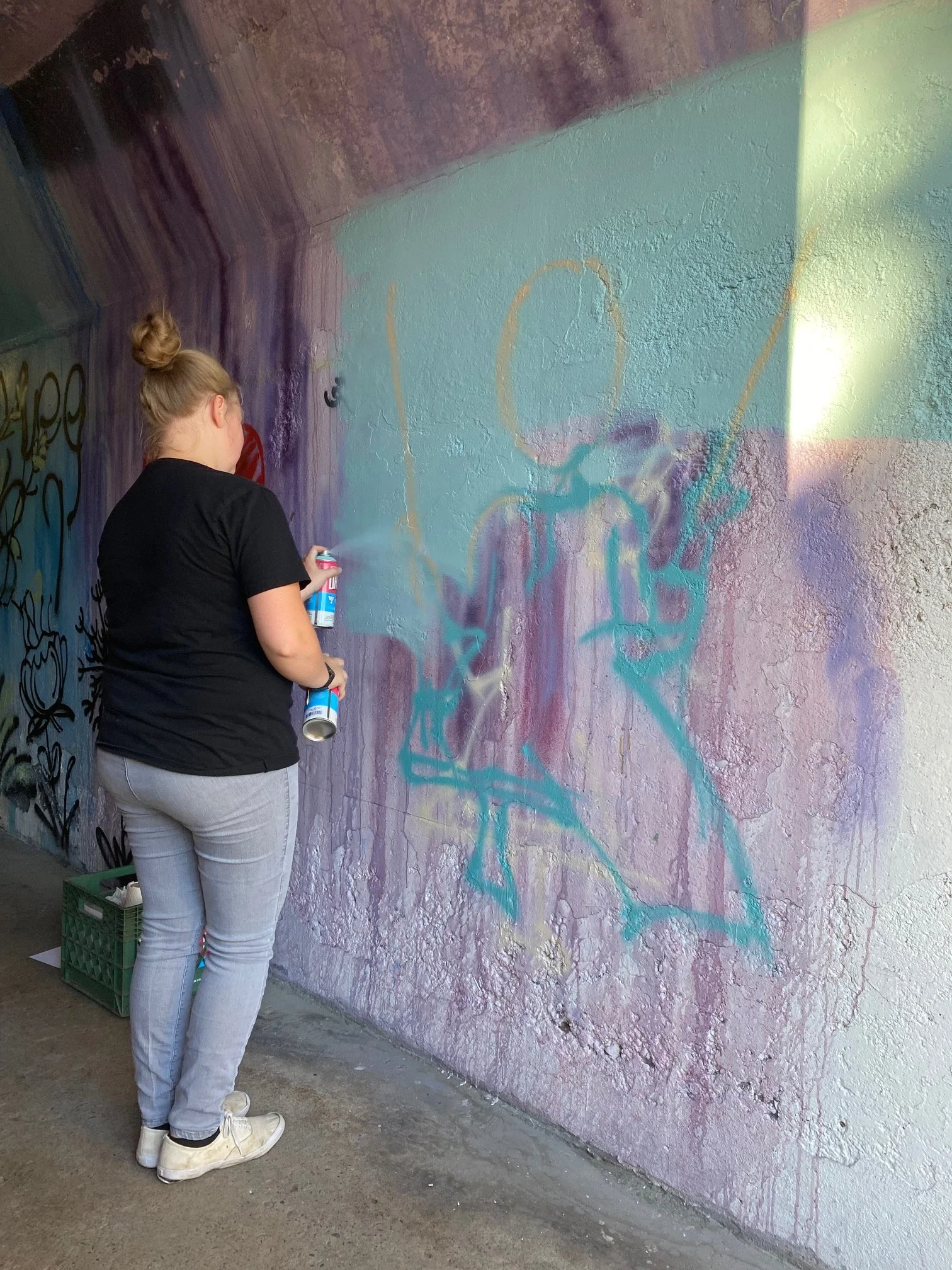 A woman with blonde hair in a bun, wearing a black t-shirt, light gray pants, and white sneakers, spray-painting graffiti on a textured wall with pink, purple, and mint colors. She is facing away from the camera, holding a spray can in her right hand