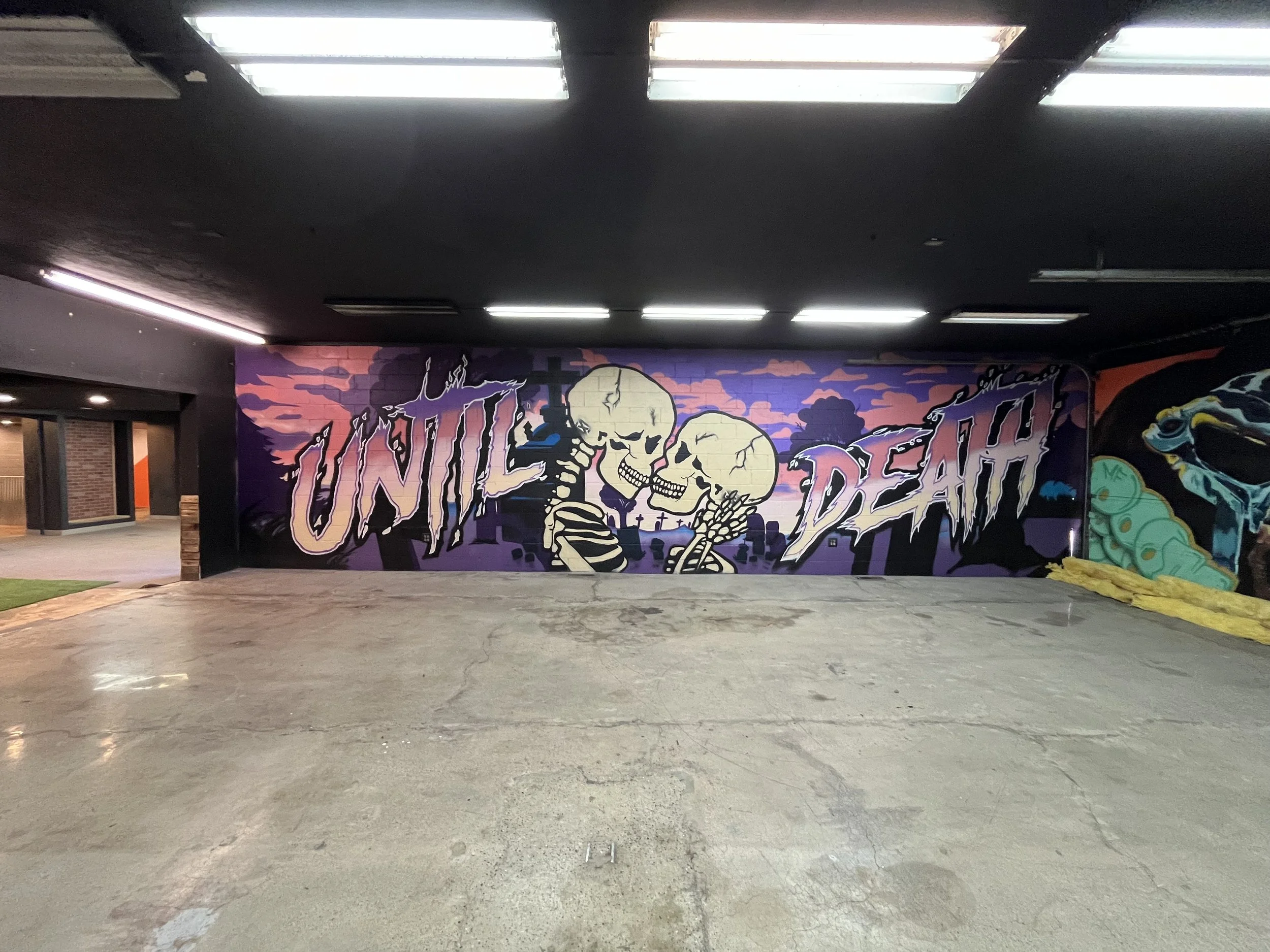 Dark indoor space with colorful graffiti art of skeletal figures and the words "Undead Death" on the wall. Fluorescent ceiling lights and an open doorway are visible.