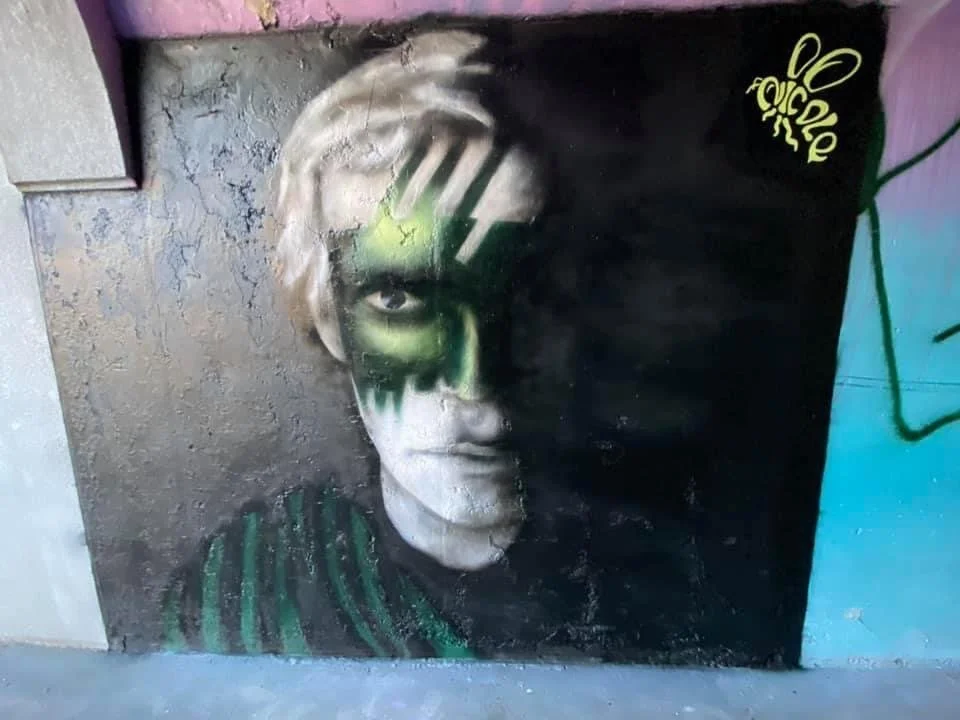 Graffiti mural of a person's face painted on a wall, with half of the face in black and white and the other half in green, with a yellow bee symbol and the words 'Bee Yourself' in yellow on the black side.