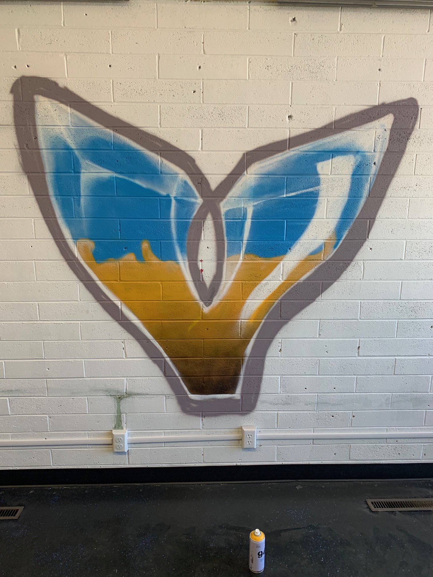 A painted mural of a butterfly on a brick wall. The butterfly has blue and yellow wings. An orange spray paint can is on the floor in front of the wall.