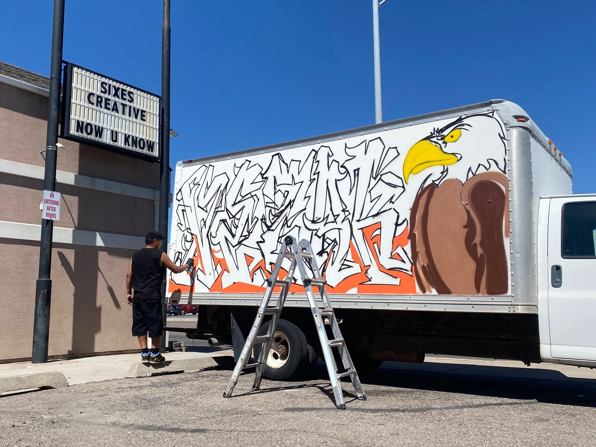A man is working on graffiti art on the side of a truck, with a ladder and tools nearby, under a clear blue sky.