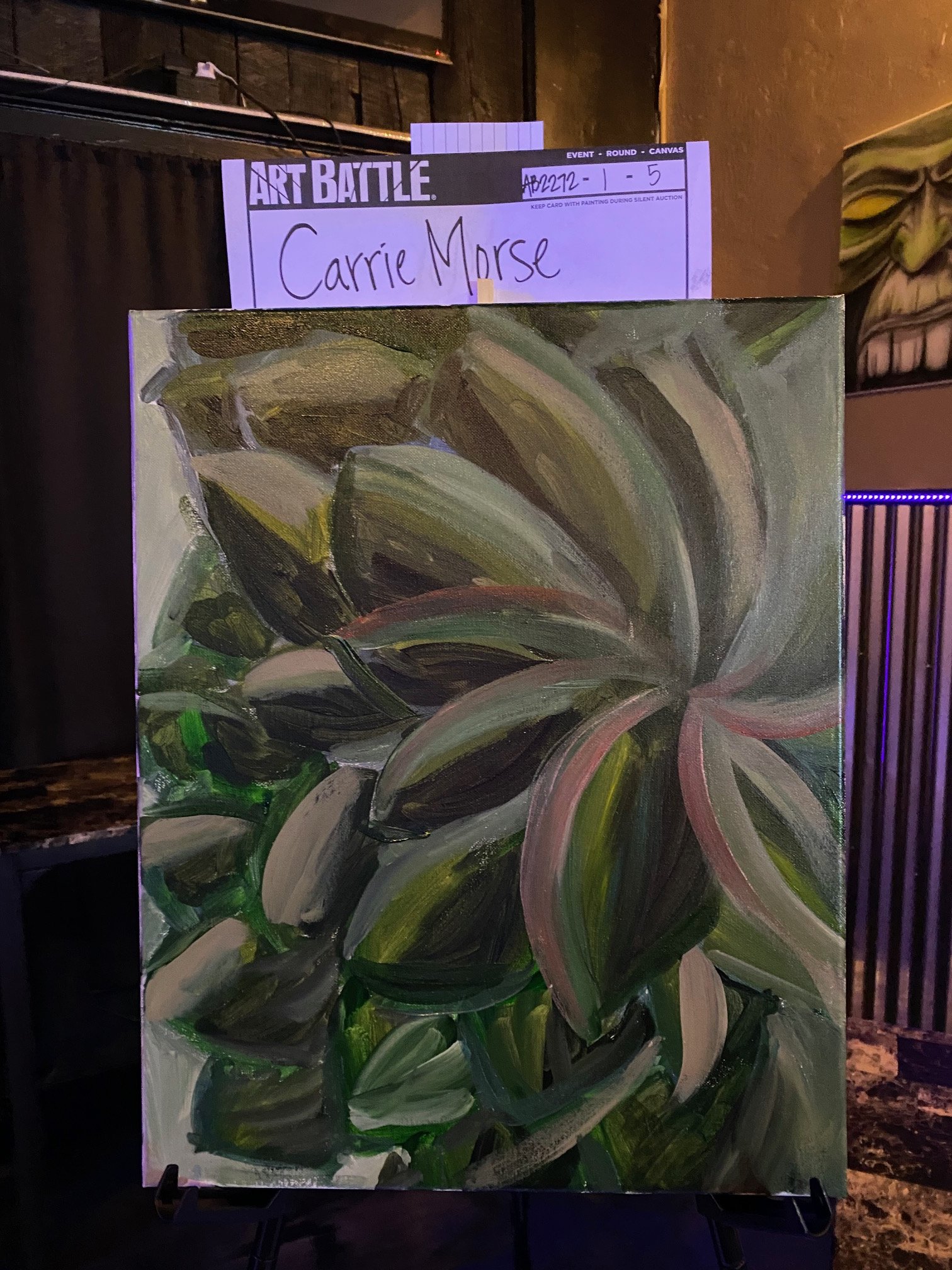 A colorful abstract painting with swirling strokes of green, black, white, and pink hues. The painting is on an easel, in an art studio or gallery setting, with a purple name tag labeled 'Carrie Morse' and a card labeled 'ART BATTLE' in the backgroun