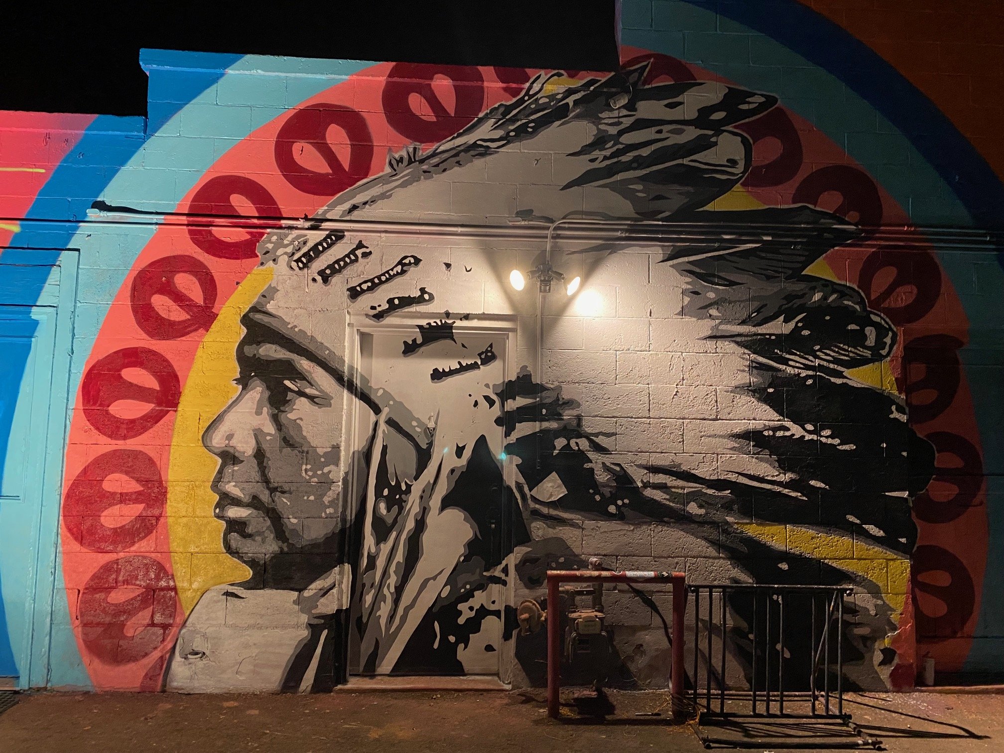 Nighttime mural of a woman with long hair, painted on a brick wall, with a rainbow and red pattern in the background.