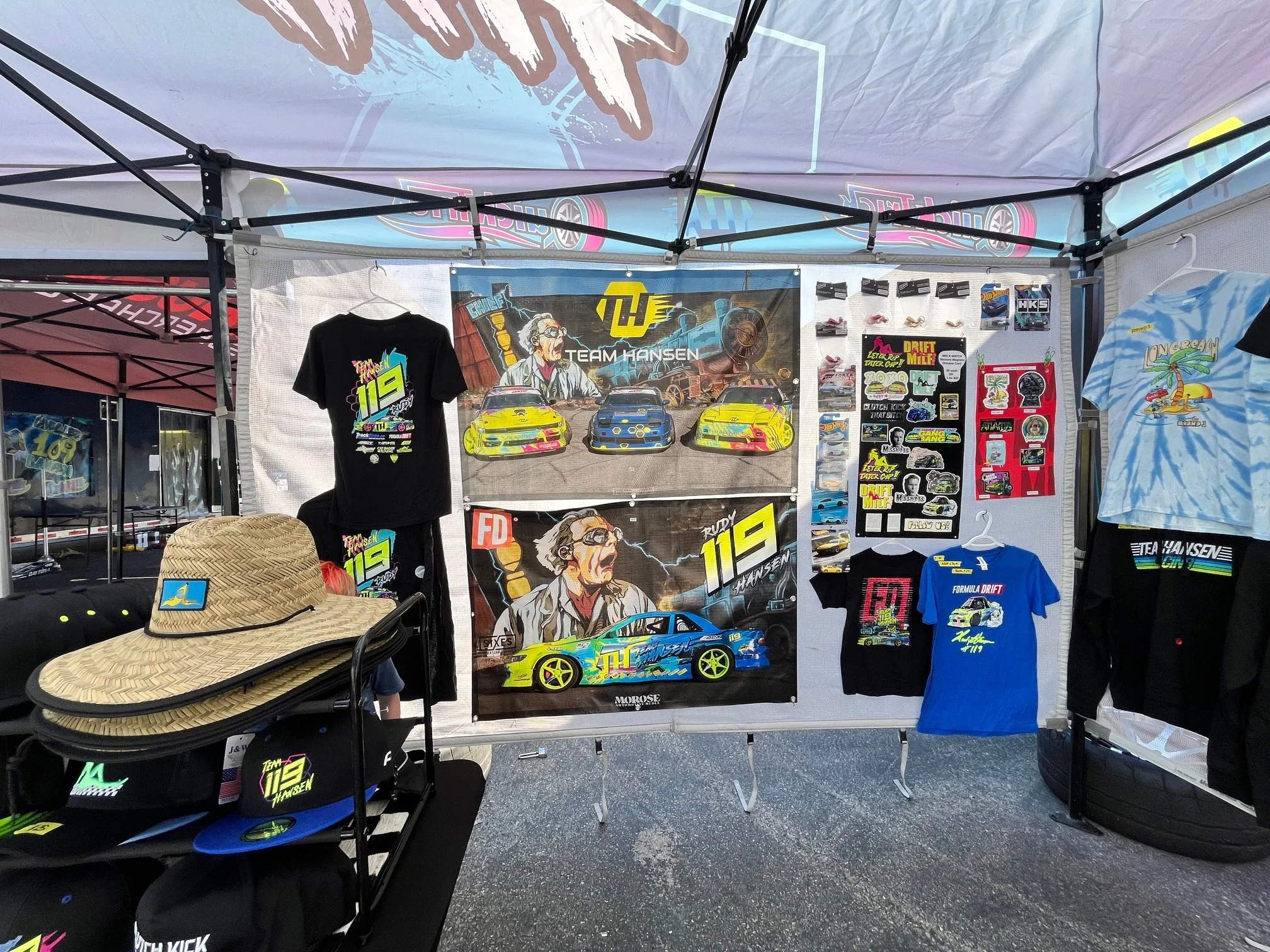 A display booth featuring racing-themed merchandise including T-shirts, posters, and accessories, with a backdrop of a racing scene and featuring a character with gray hair and glasses. On the table, a large straw sun hat and small model cars are vis