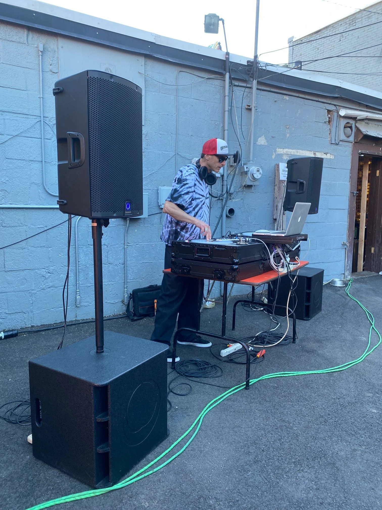 A person in a tie-dye shirt, sunglasses, and a red cap with white front, DJing outdoors with audio equipment, two large speakers, and cables, against a light blue brick wall.