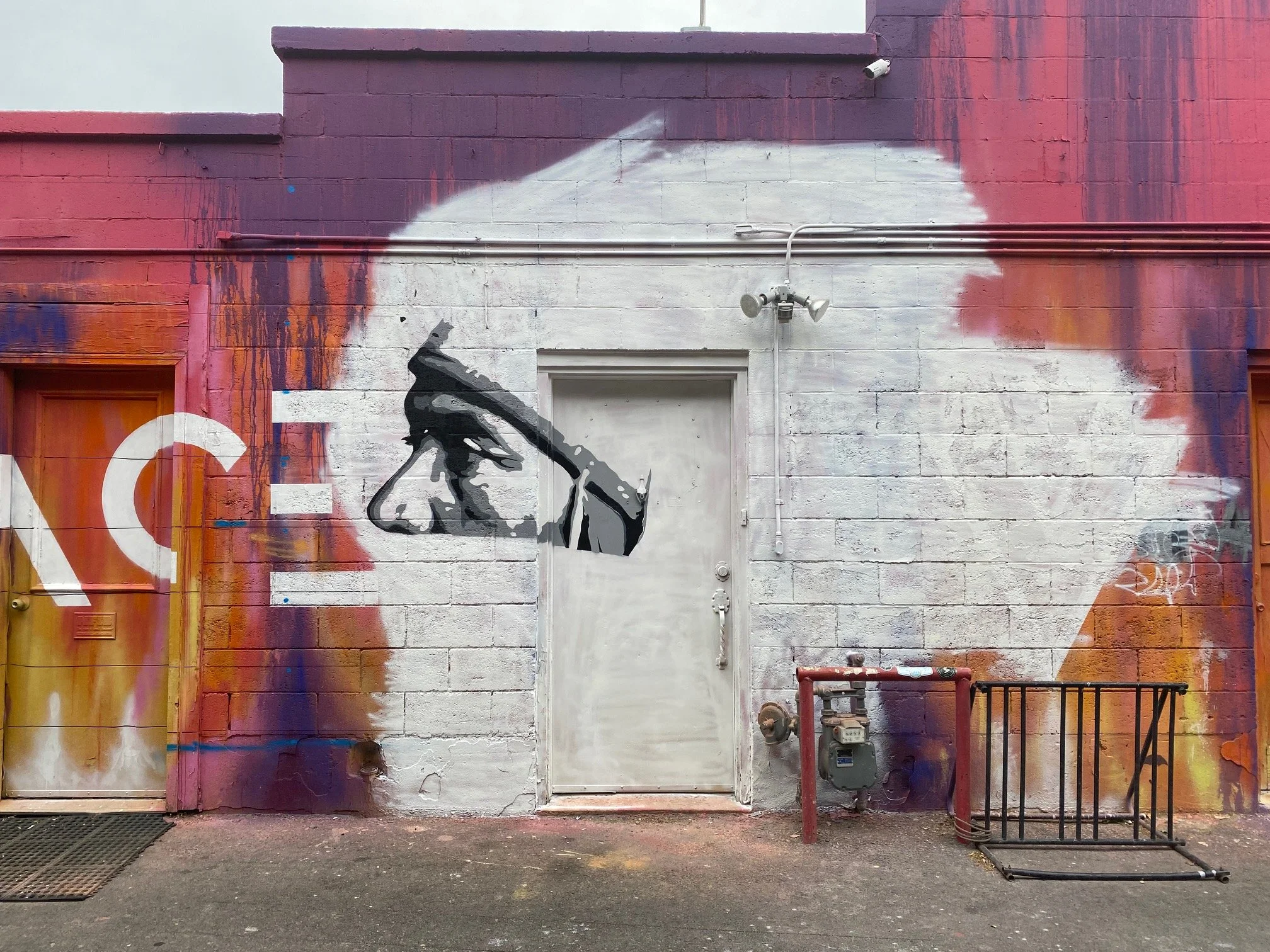 Street art mural on a brick wall featuring a black and white profile of a person's face with their hand covering part of their face and a large white cloud shape painted behind them. The wall includes electrical wiring and security cameras.