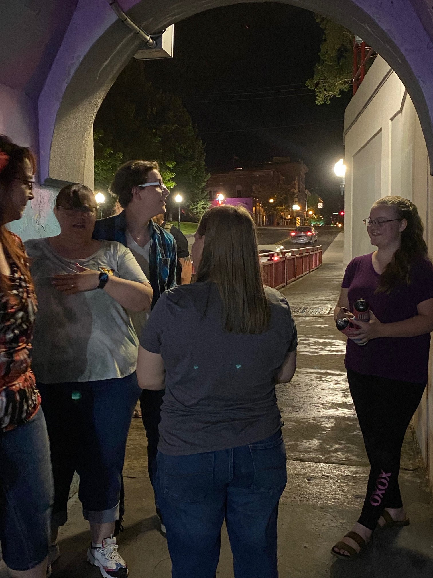 Group of six women standing and talking under an archway at night, with city street lights and buildings visible in the background.