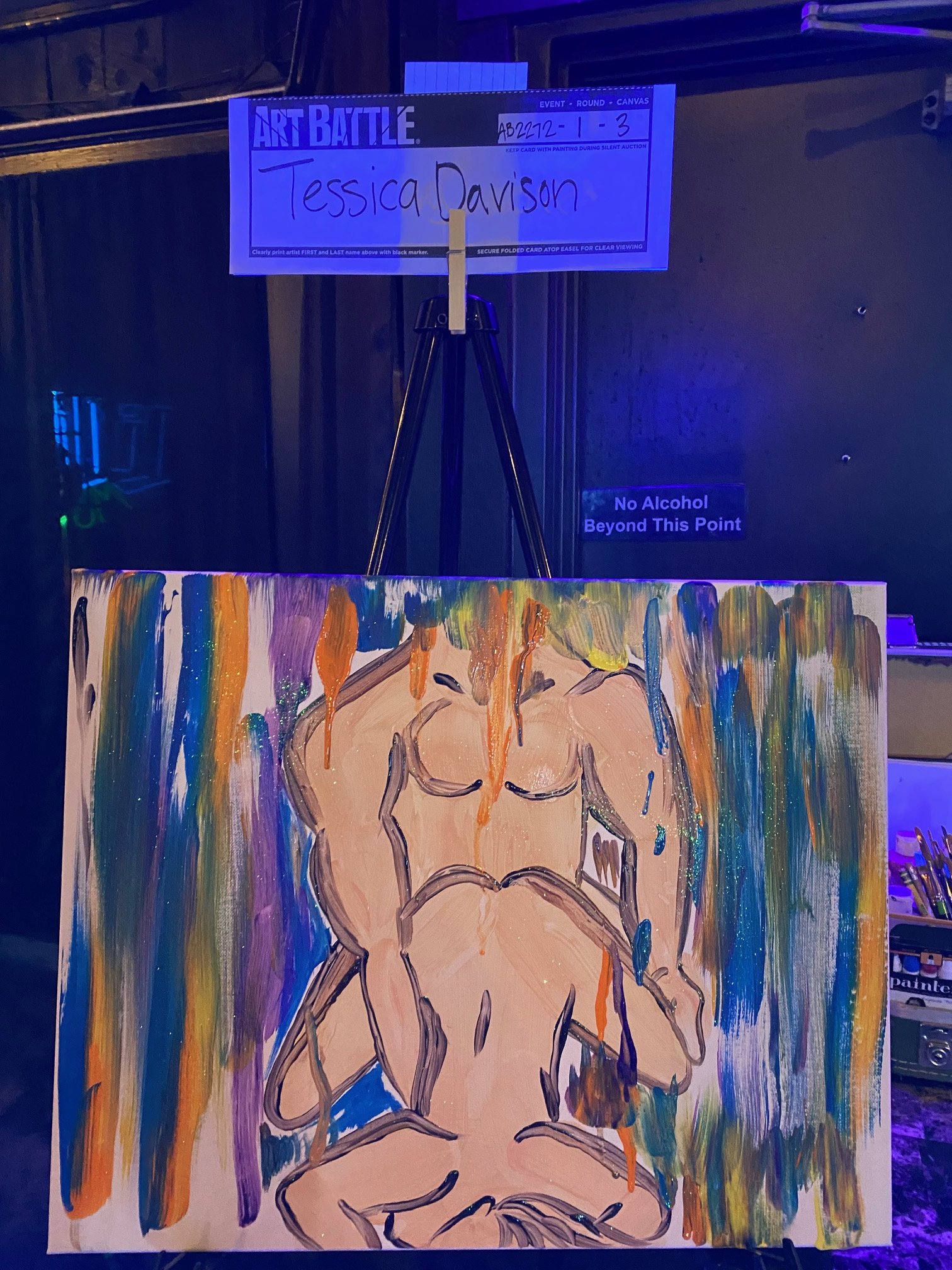 An art piece on a canvas showing a nude figure painted with rainbow colors and drips, with a sign in the background indicating an art battle event for artist Tessica Davison.