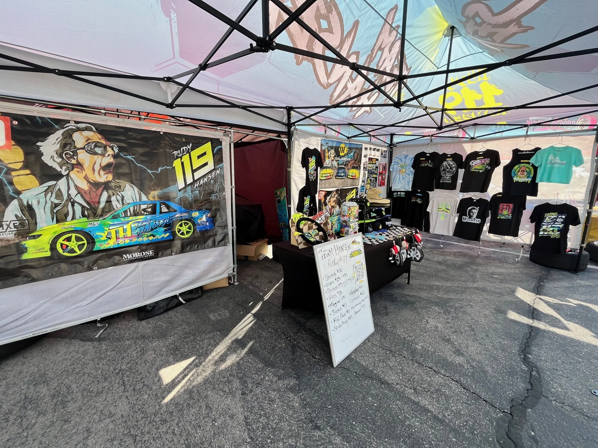 A vendor booth at an outdoor event with a large banner of a cartoon character wearing sunglasses and a racing suit. The booth sells various racing-themed merchandise, including t-shirts, hats, and toy cars. A whiteboard displays prices for different 