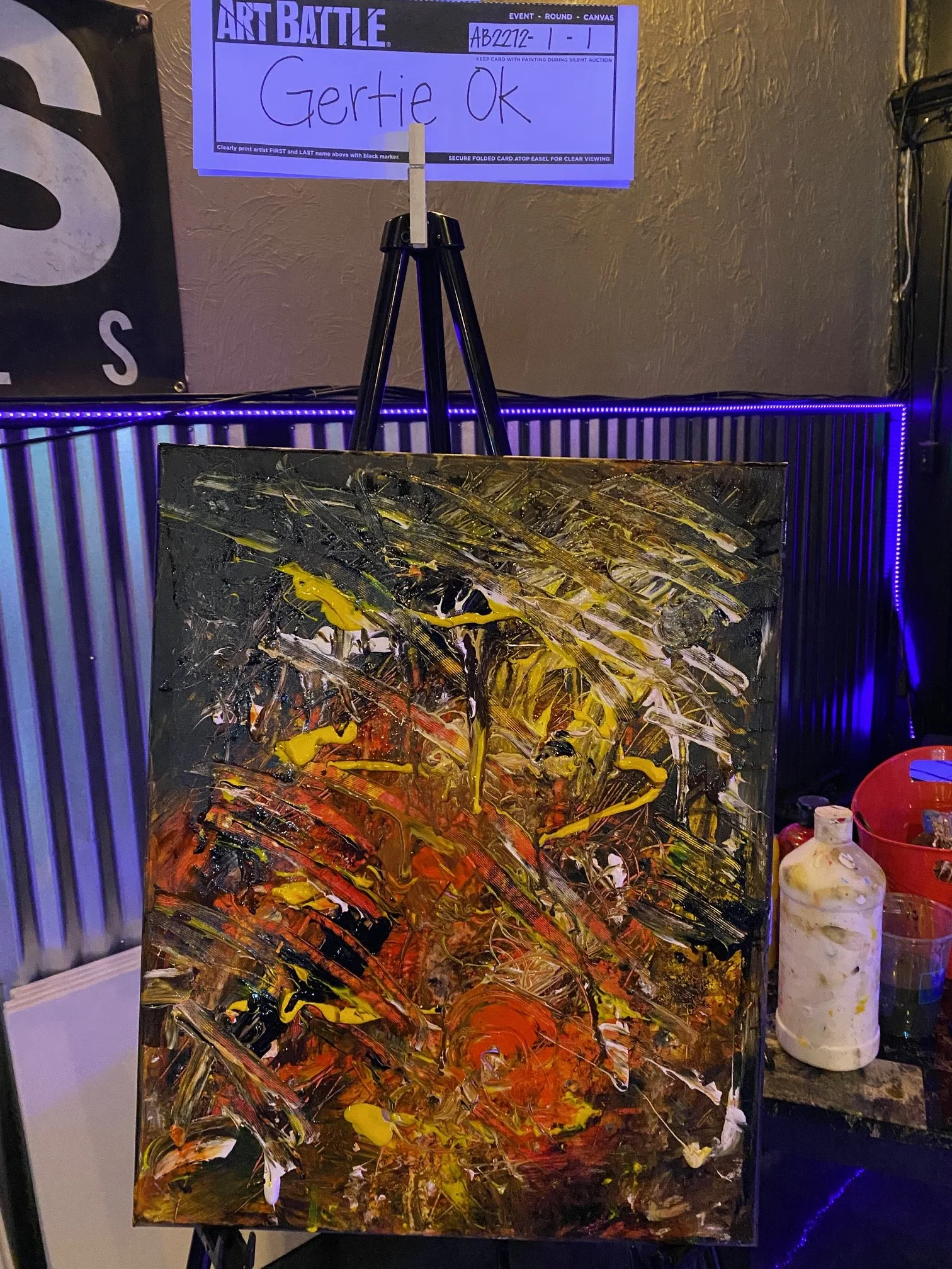 Abstract expressionist painting with bold strokes of yellow, orange, black, red, and white paint on canvas, displayed on an easel in an art gallery. A sign above indicates this is part of an art battle event, with the name "Gertie Ok" written on the 