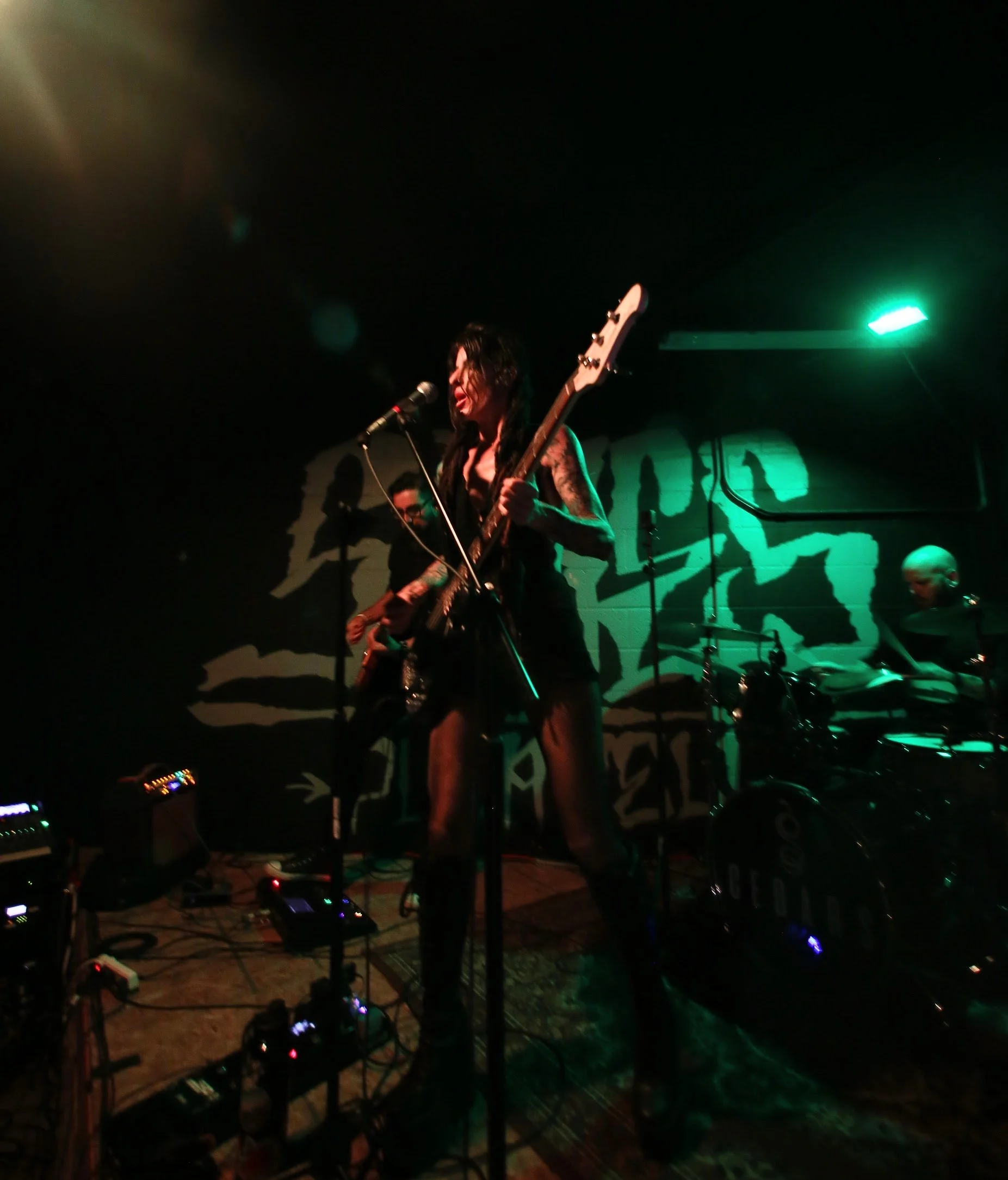 Female musician singing and playing electric guitar on stage in a dark venue, with drummer in background and green stage lighting.