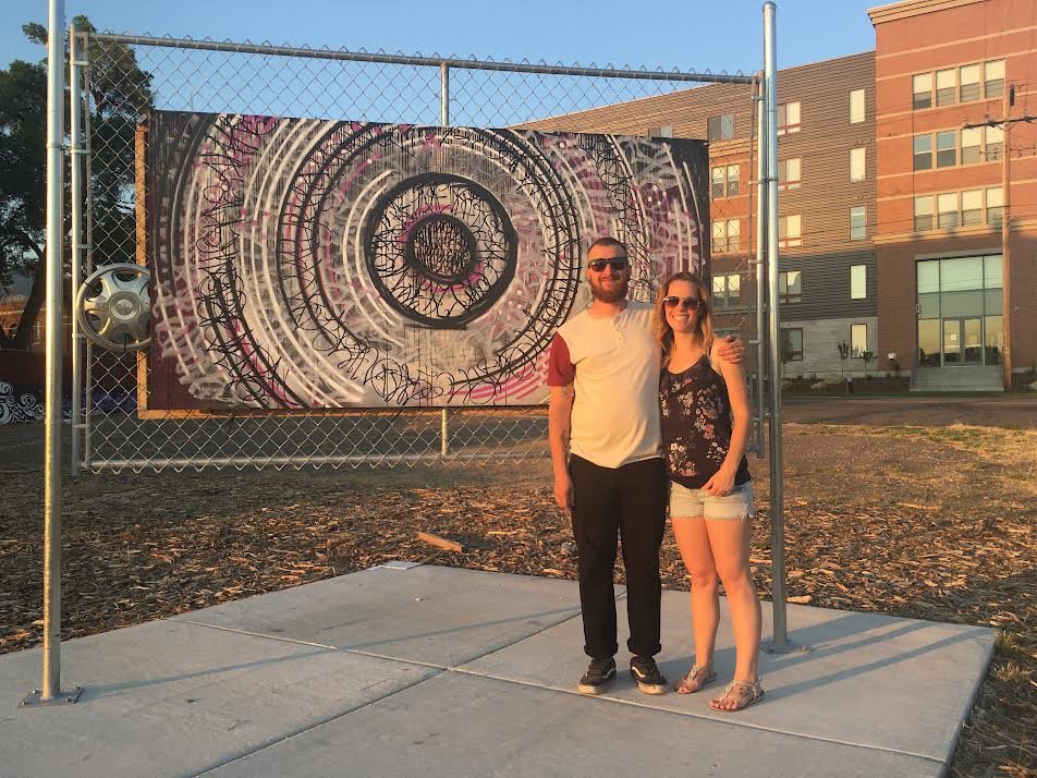 A man and woman standing on a sidewalk in front of an artistic graffiti mural on a chain-link fence, with a residential building in the background.