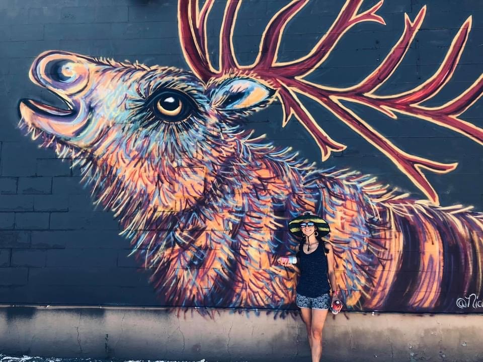 A woman wearing a hat and sunglasses standing in front of a large, colorful mural of a deer on a brick wall