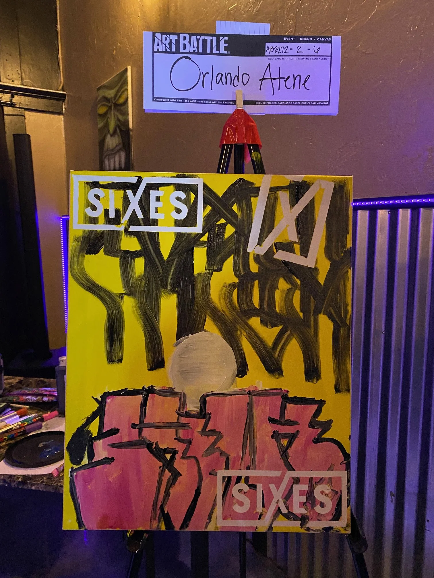 Colorful paintings on an art display board with the words 'SIXES' and other artwork, under a sign that reads 'ART BATTLE'.
