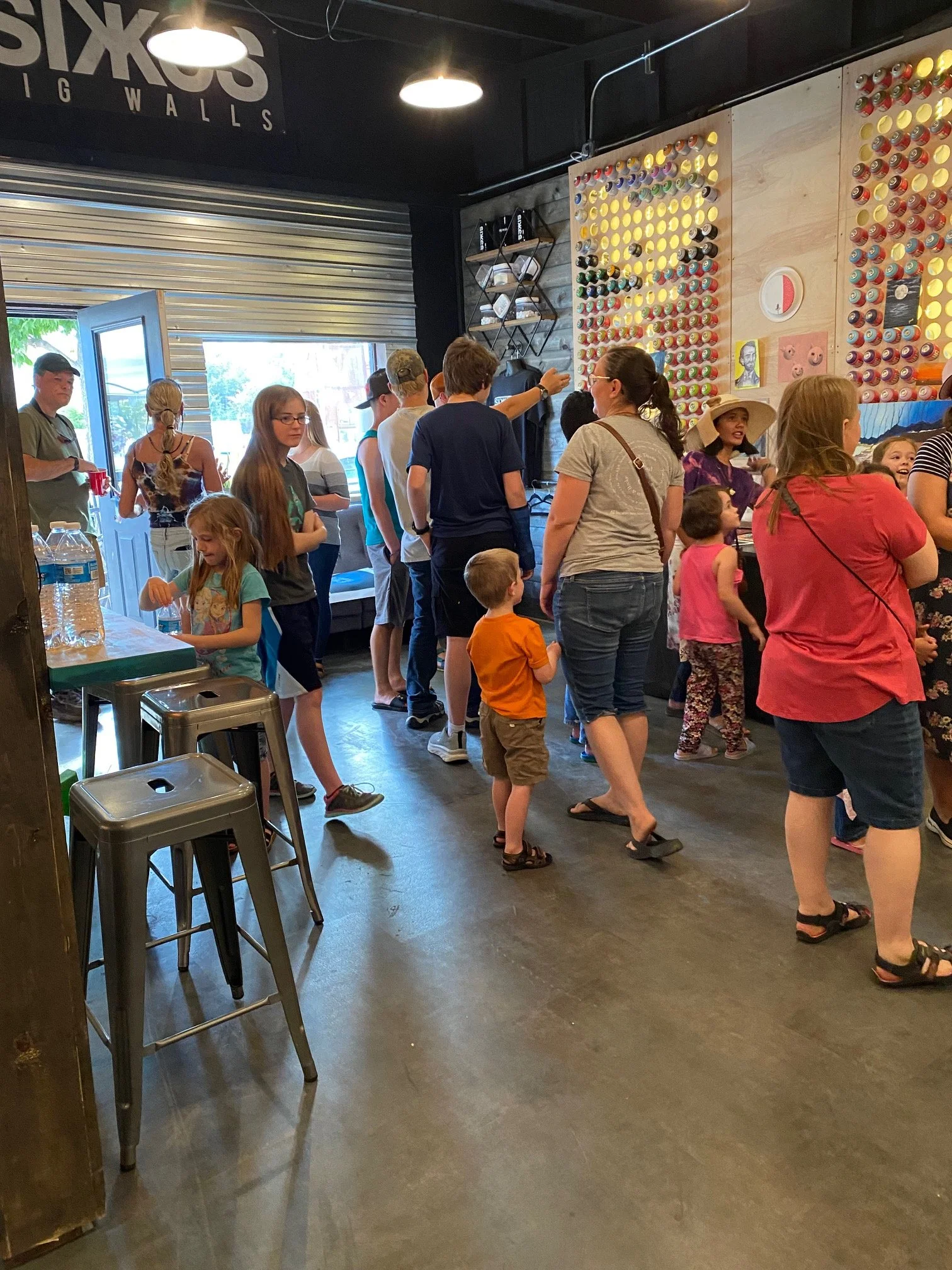 A group of people, including children and adults, standing inside a tattoo shop, looking at wall art and waiting, with some seated near the entrance. The shop has a wall decorated with numerous spray paint cans, and a black and wooden interior with s