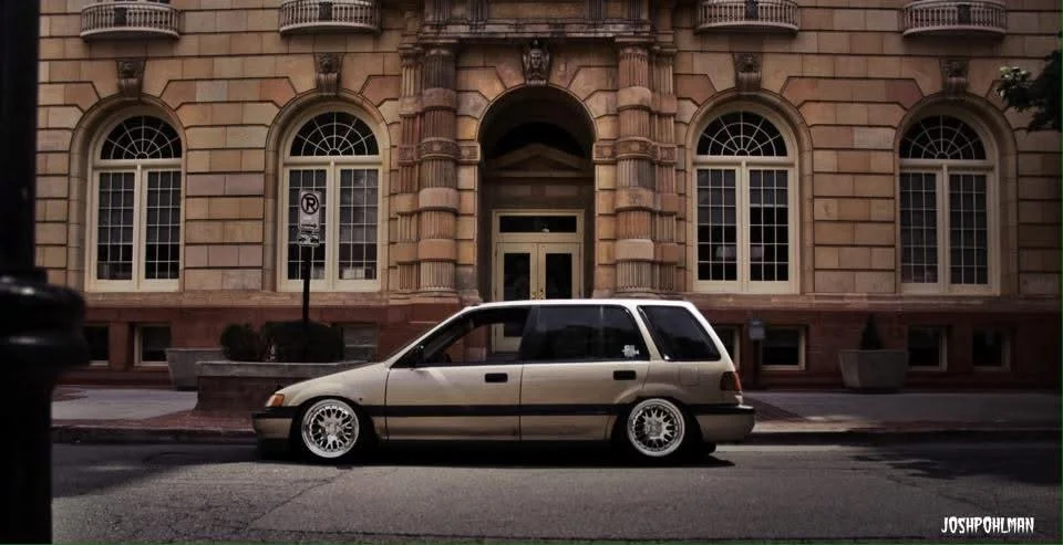 A beige compact car with lowered suspension and white aftermarket wheels parked in front of a historic stone building with arched windows and decorative columns.