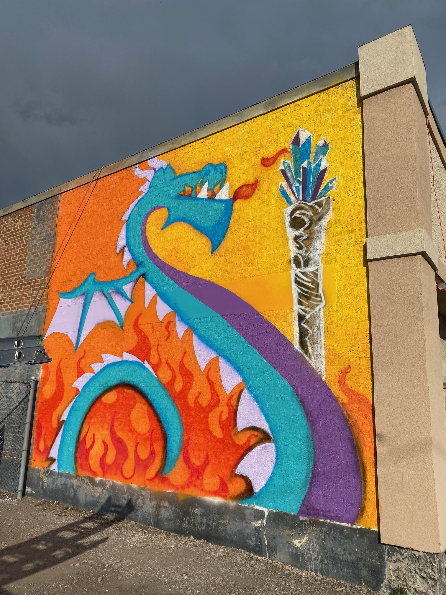 A colorful mural on an outdoor brick wall features a blue dragon and a silver cup filled with multi-colored crystals, set against a yellow and purple background with fiery orange accents.
