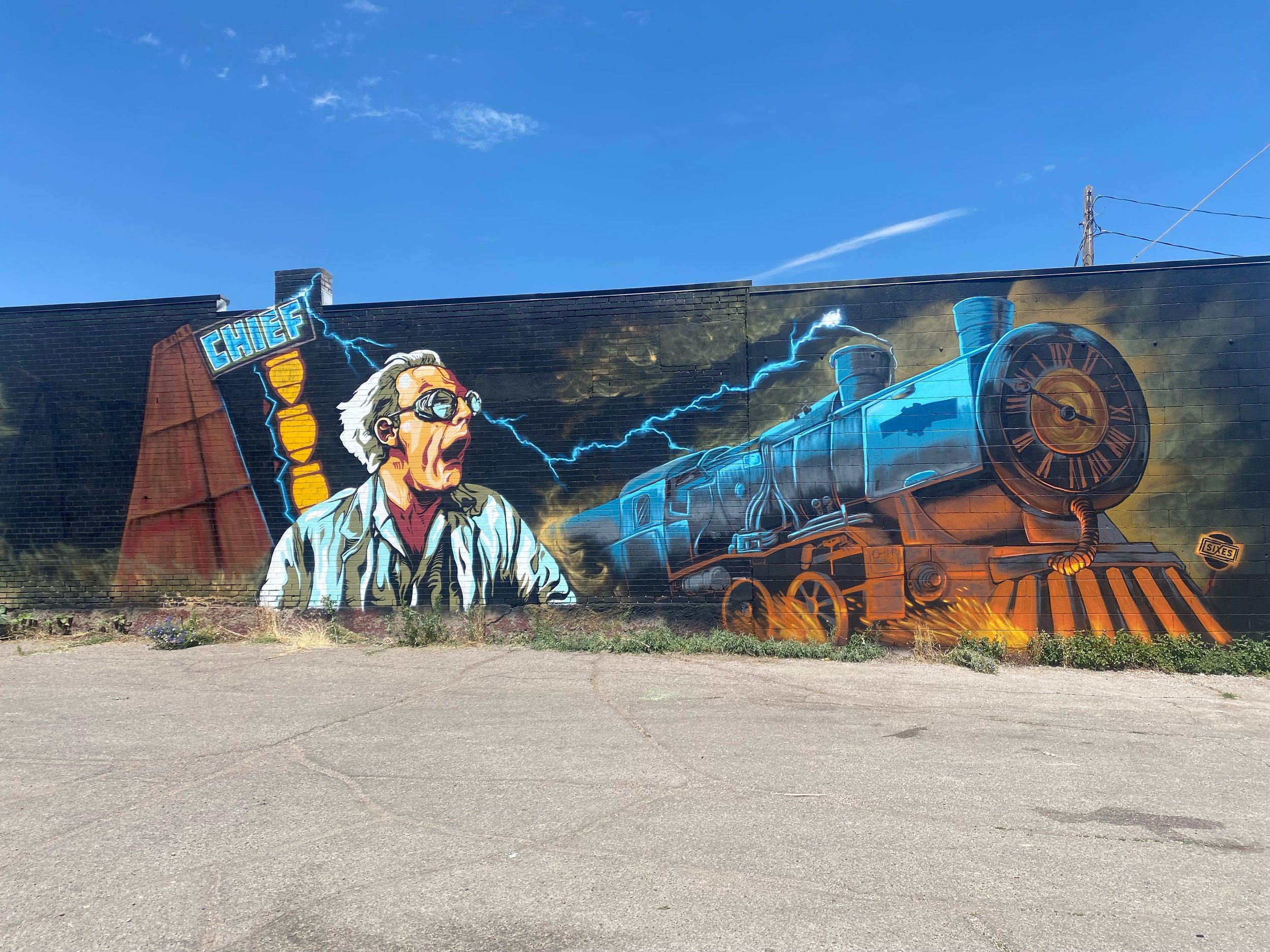 Colorful mural on a brick wall depicting a scientist with grey hair and glasses, a large mechanical train emitting electricity, and a red building with a sign that says 'CHIEF' under a blue sky with a few clouds.