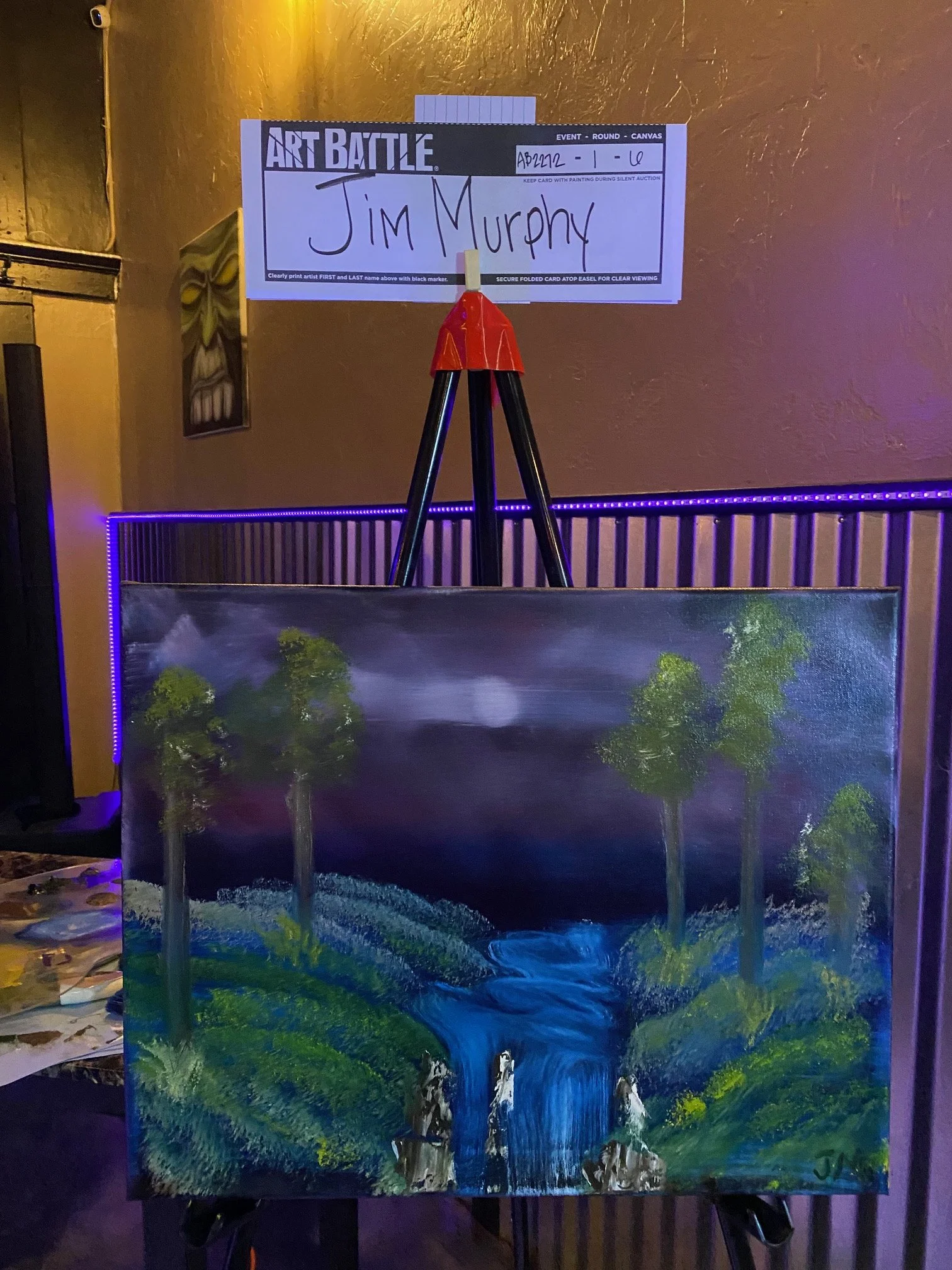 A painting of a waterfall flowing into a river, surrounded by trees and greenery, on an easel in an art gallery, with a sign for an art competition or event above it.
