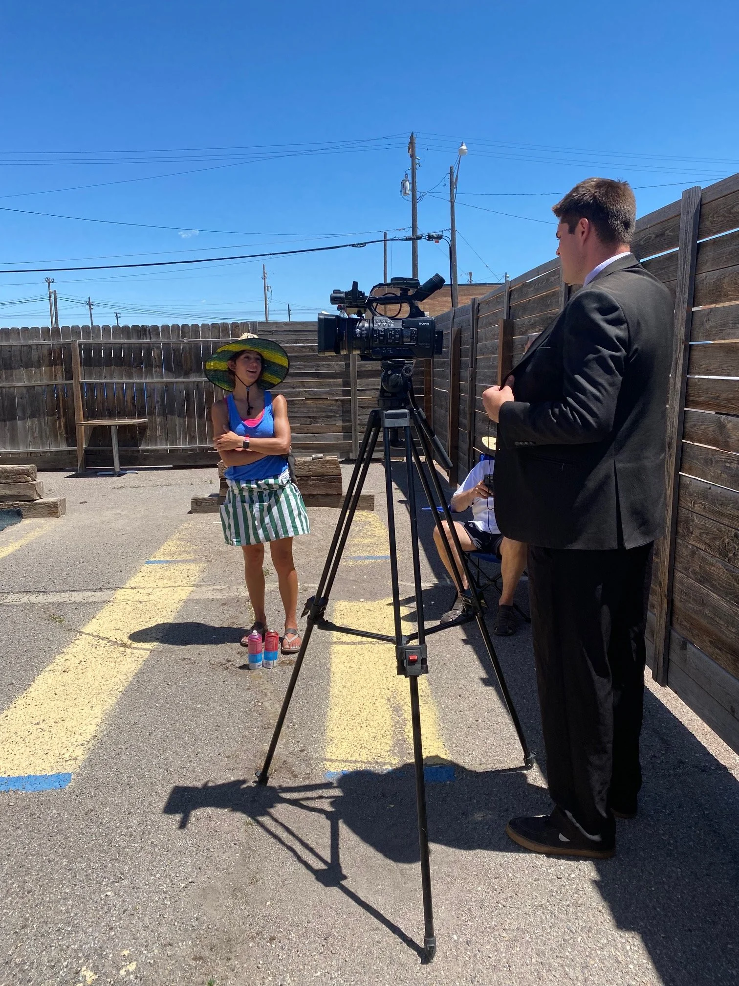 A woman in a blue tank top, striped skirt, and large straw hat stands outdoors with her arms crossed, smiling and talking to a man in a black suit, who is standing in front of a professional video camera. The scene is set in a parking lot with a wood