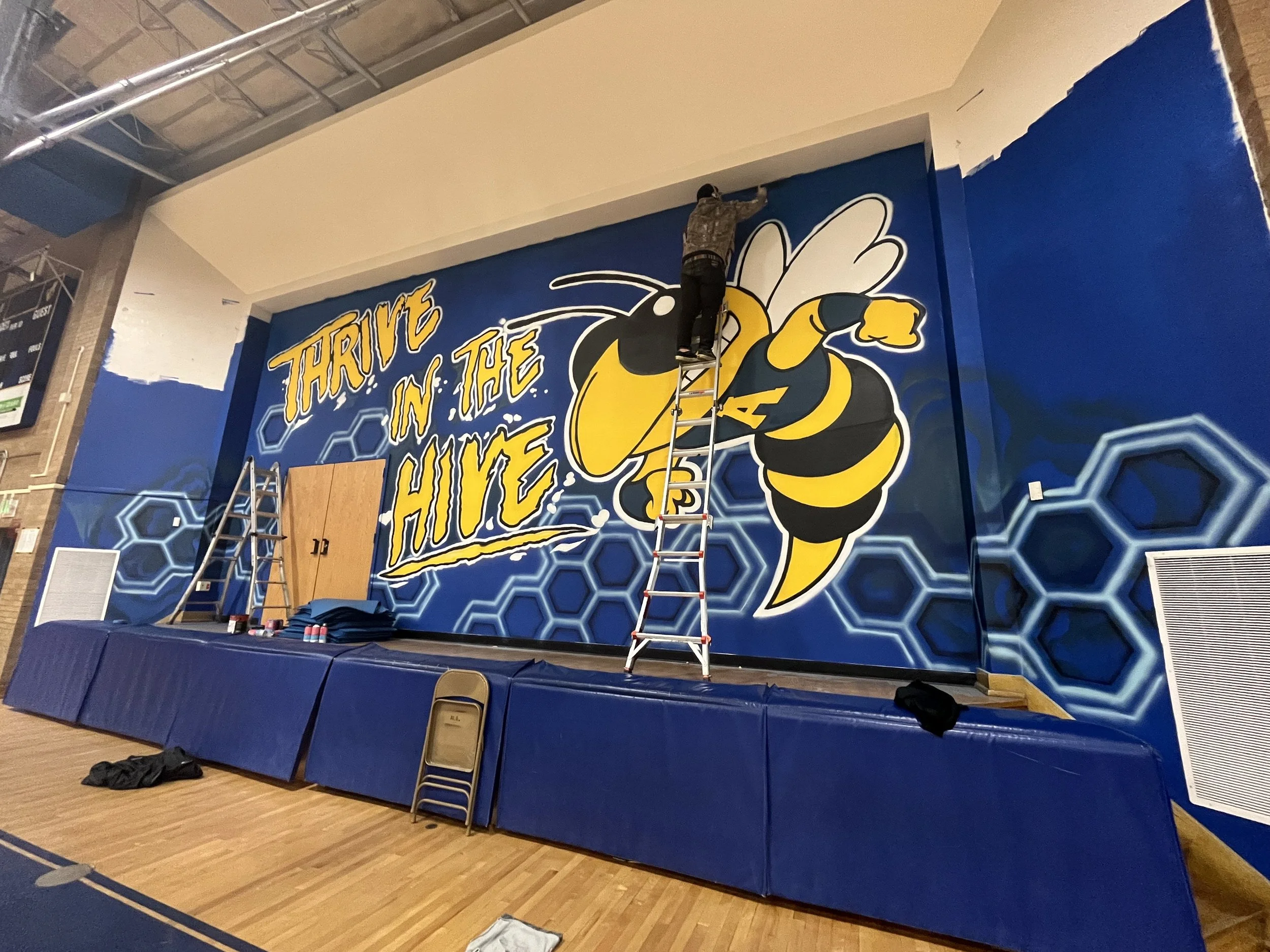 A person is standing on a ladder painting a large wall mural of a bee with the phrase "Thrive in the Hive" in a gymnasium or indoor sports facility.
