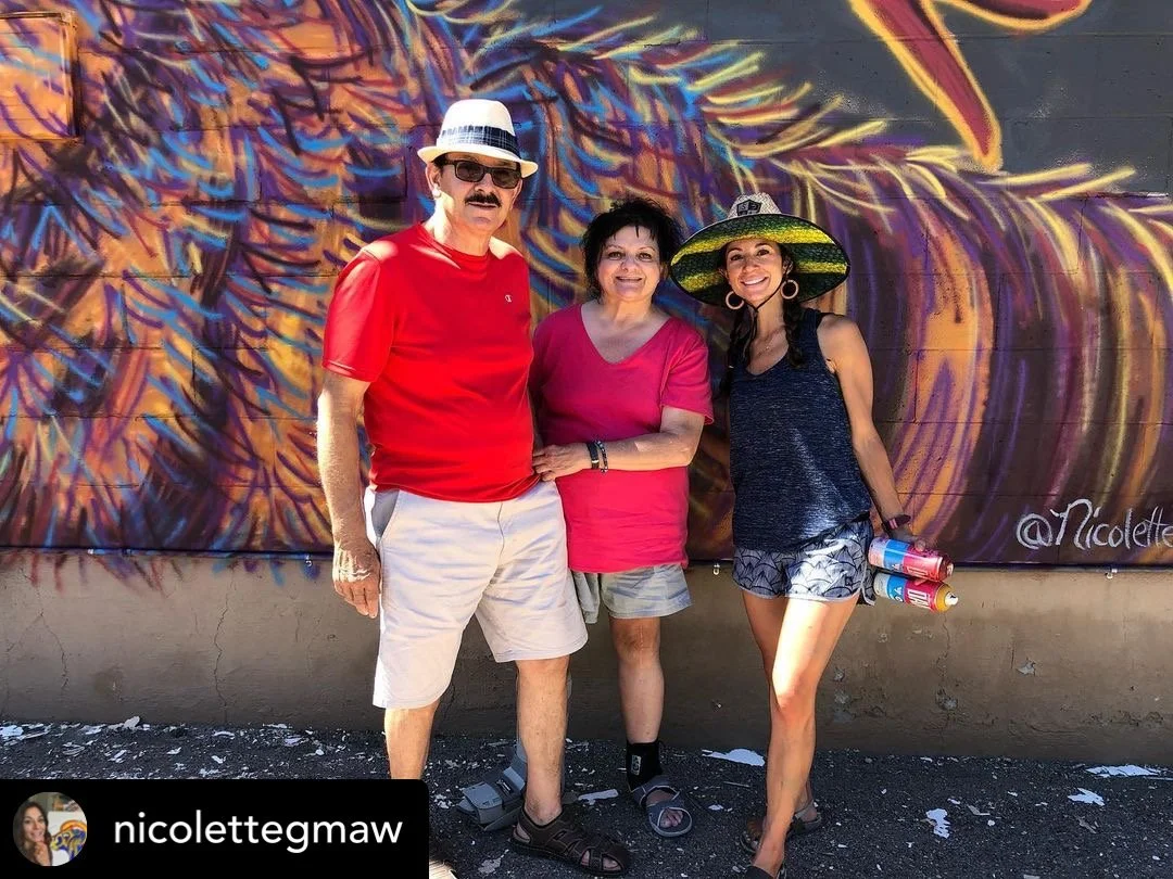 Three people standing in front of a colorful mural. The man on the left is wearing a red t-shirt, beige shorts, hat, sunglasses, and sandals. The woman in the middle is wearing a pink t-shirt, patterned shorts, and sandals. The woman on the right is 