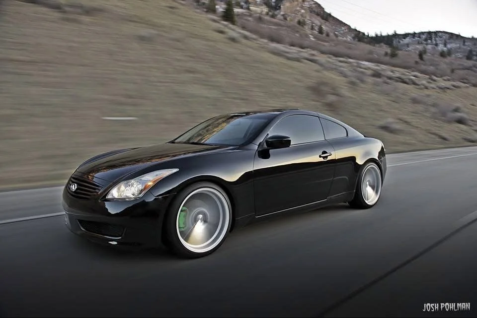 A black Infiniti coupe driving on a highway with a mountainous landscape in the background.