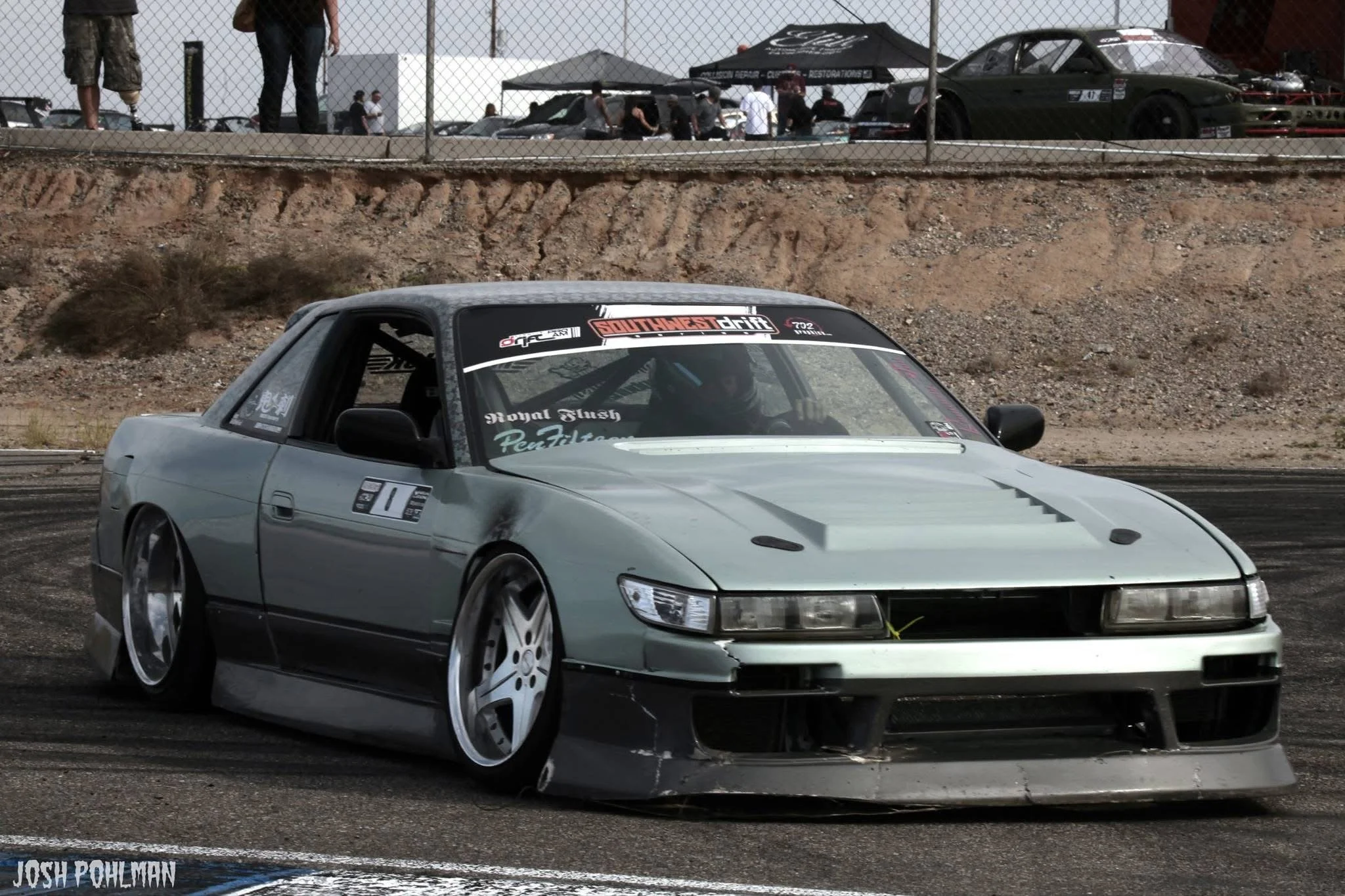 A silver sports car with modified wheels and lowered suspension on a racetrack, with a bandage near the front wheel and a sticker on the front bumper reading "Josh Pohlman." In the background, there are people near a chain-link fence and several othe