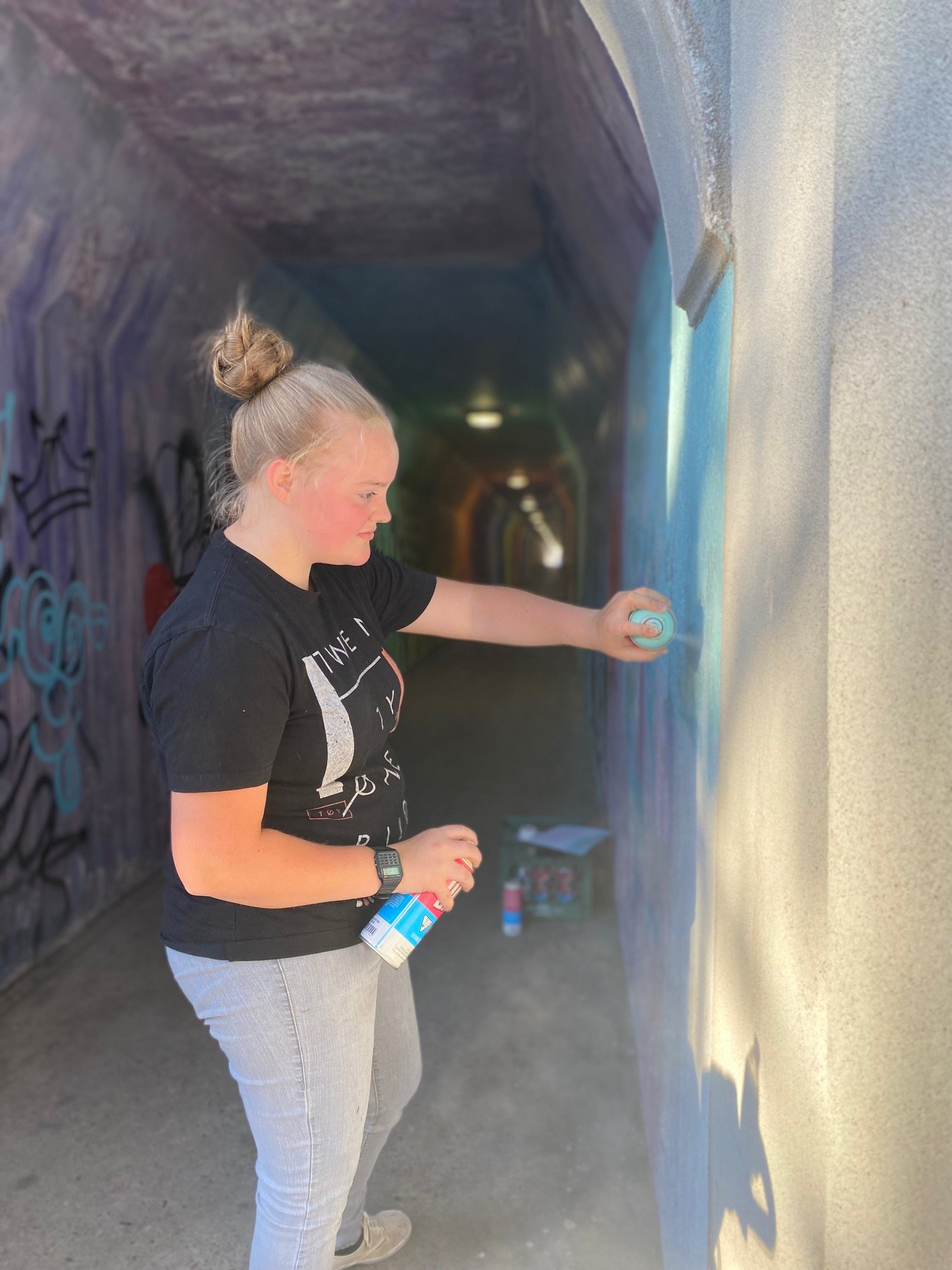 A woman with a bun hairstyle and gray pants is spray painting graffiti on a wall inside a tunnel. She is holding spray paint cans and is wearing a black T-shirt with writing on it.