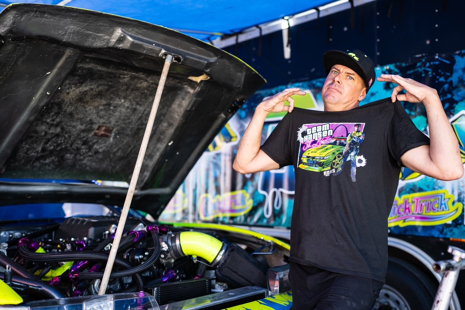A man wearing a black t-shirt with race car graphic and a black cap stands next to a racing car with its hood open, showing the engine and mechanical parts, in a racing pit area.
