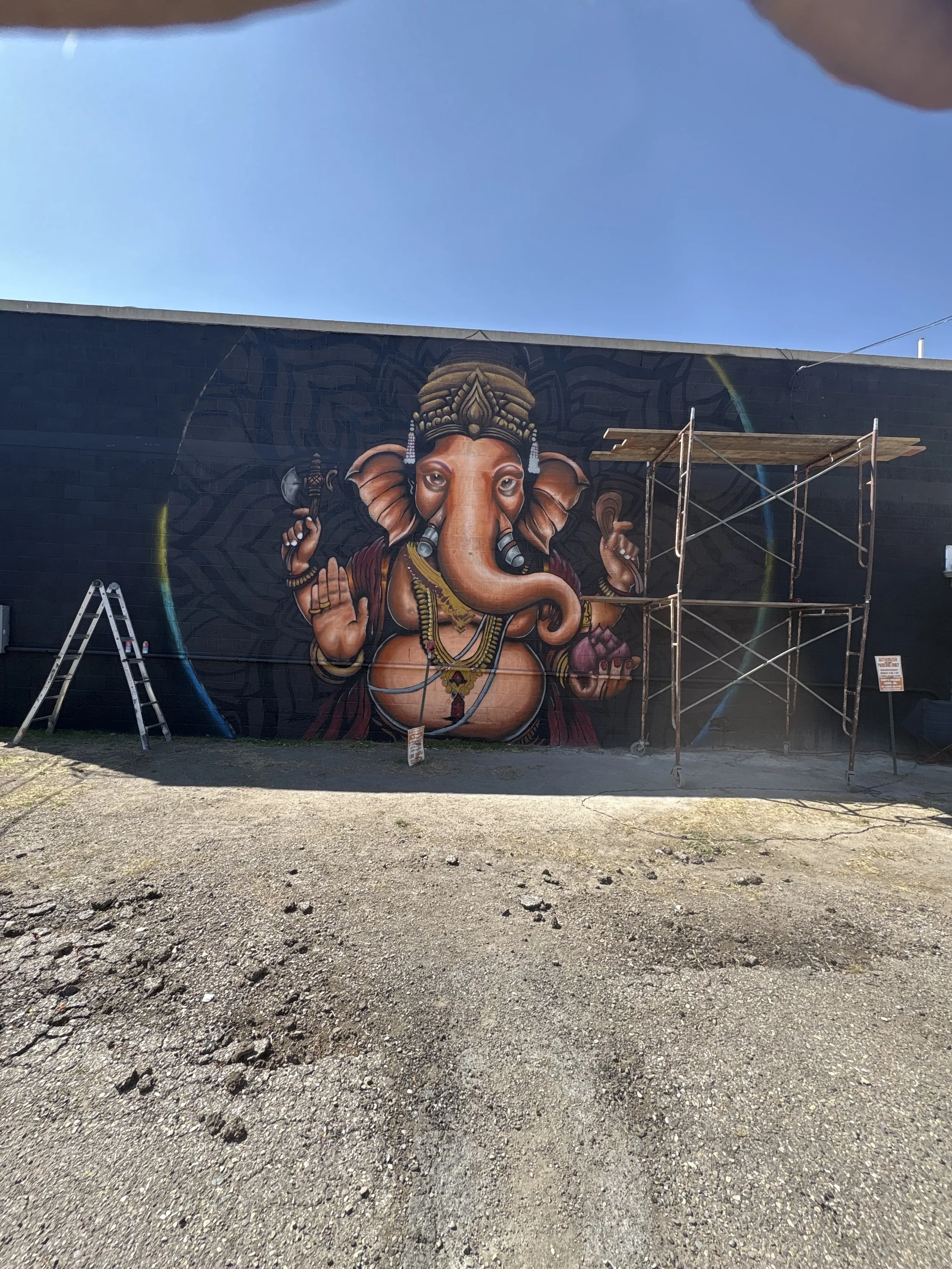 Street view of a colorful mural of the Hindu deity Ganesha painted on a black wall, with scaffolding and a ladder nearby, under a clear blue sky.