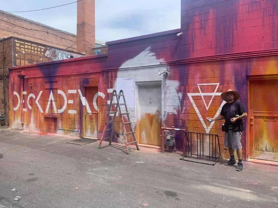 A person in a black shirt, gray shorts, sneakers, and a wide-brimmed hat stands in front of a colorful graffiti mural on a brick building. The mural features large white letters spelling 'STACKADE' and geometric shapes on a multicolored background.
