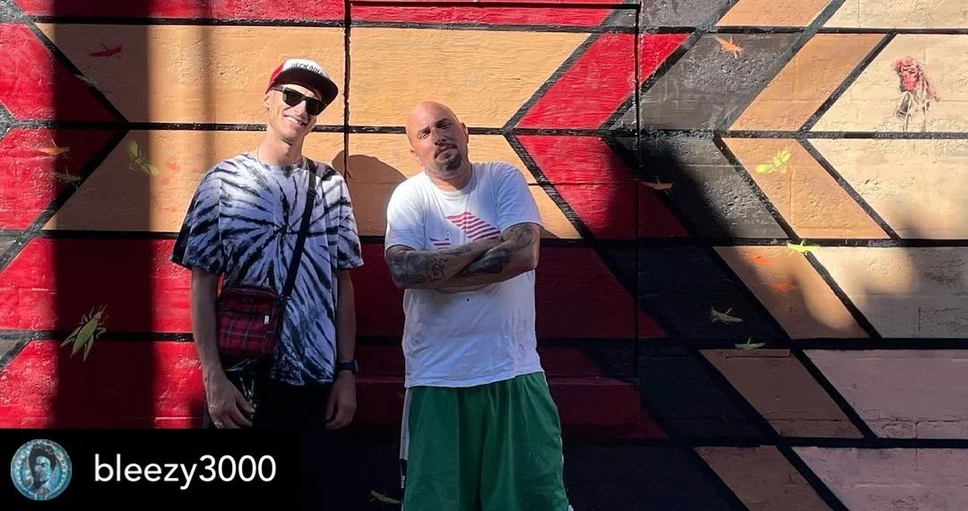 Two men standing in front of a colorful mural on a brick wall. The man on the left is wearing sunglasses, a hat, a tie-dye t-shirt, and a crossbody bag. The man on the right is bald, with tattoos on his arms, wearing a white t-shirt with an American 