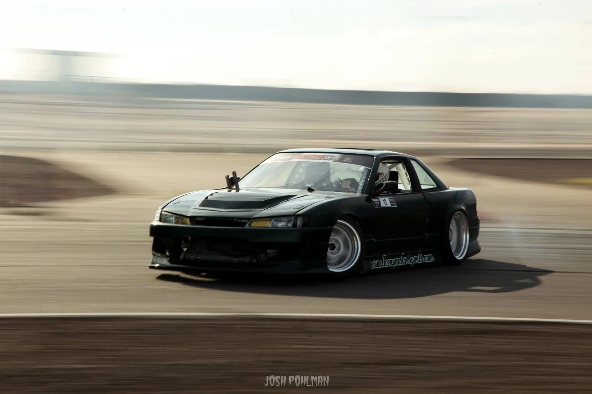 A black race car with a hood scoop and wide tires speeding on a race track, with motion blur in the background.