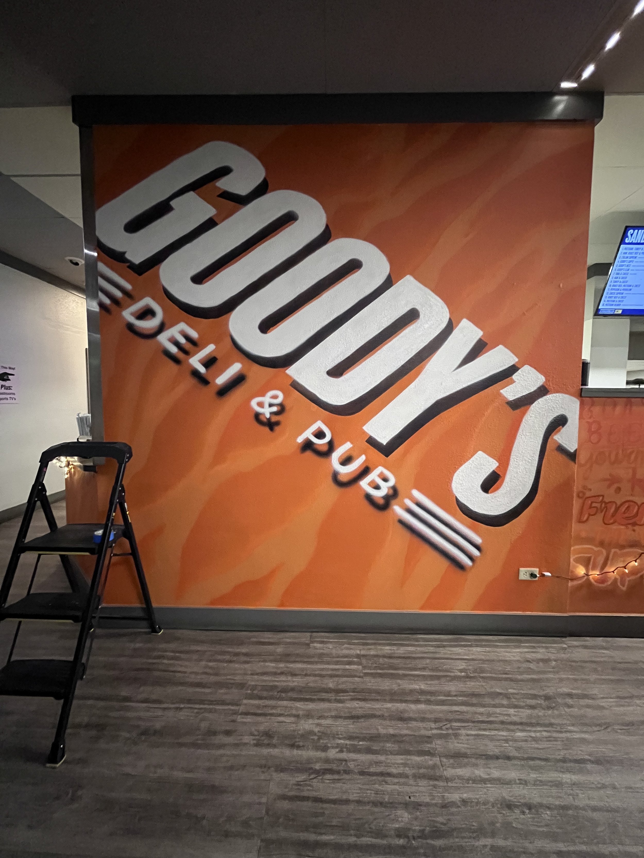 A large orange advertisement banner with white and black text promoting a food or beverage product, inside a building with a wooden floor and a small black staircase in front of it, and a television screen on the right side.