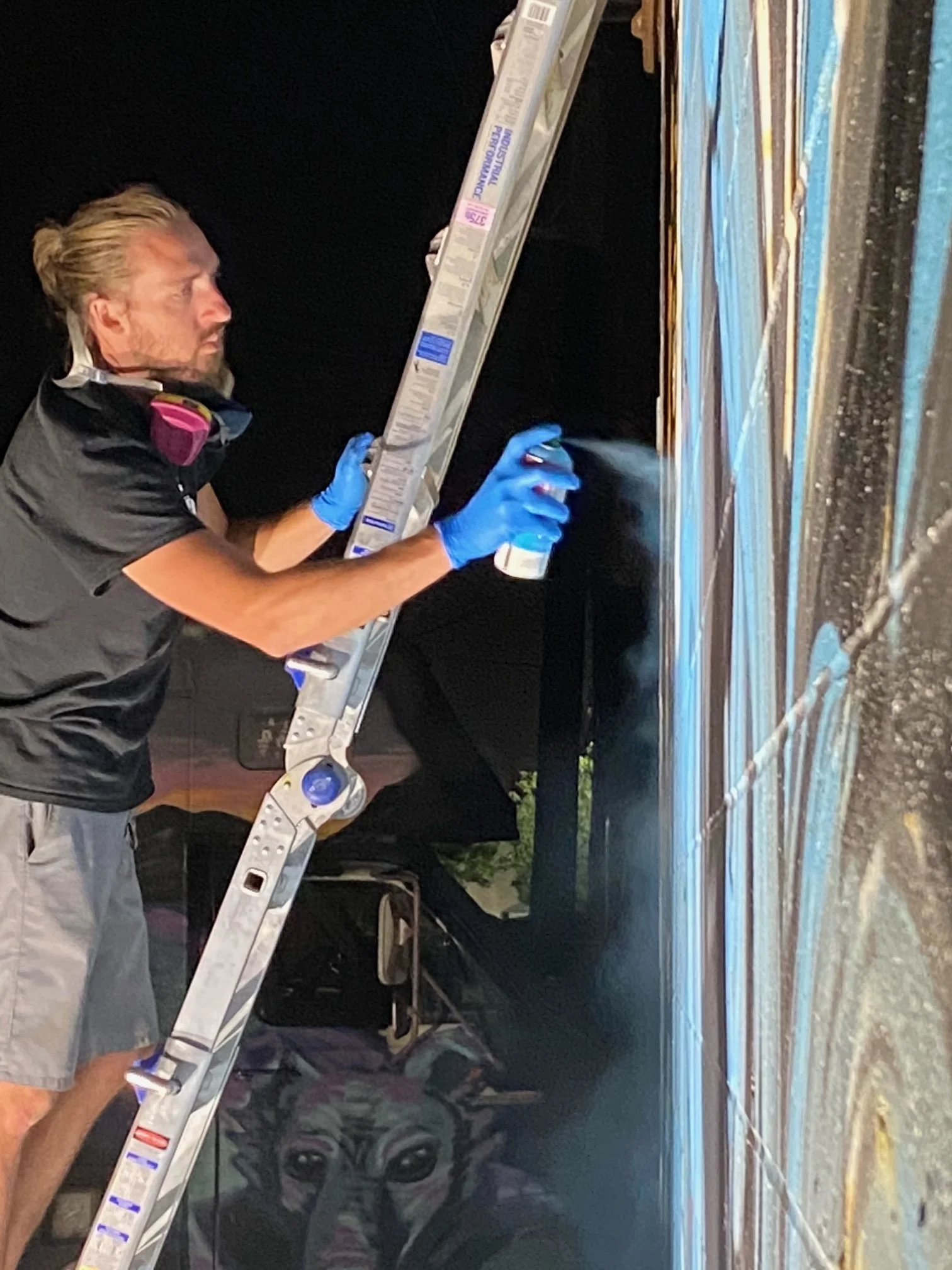 A man wearing blue gloves cleaning a surface with a spray bottle next to an aluminum ladder at night.