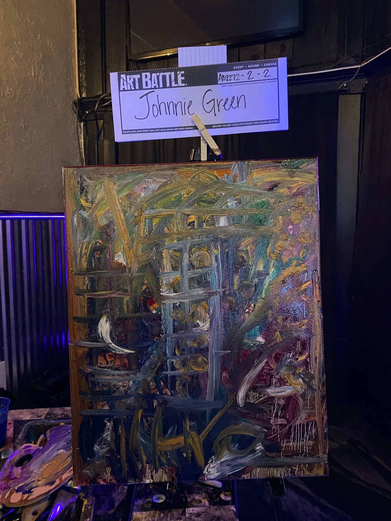 An abstract painting with bold, sweeping strokes of yellow, green, purple, and white across a canvas, set on an easel. A sign above the painting reads 'ART BATTLE' with the name 'Johnnie Green' handwritten on it, indicating participation in an art co