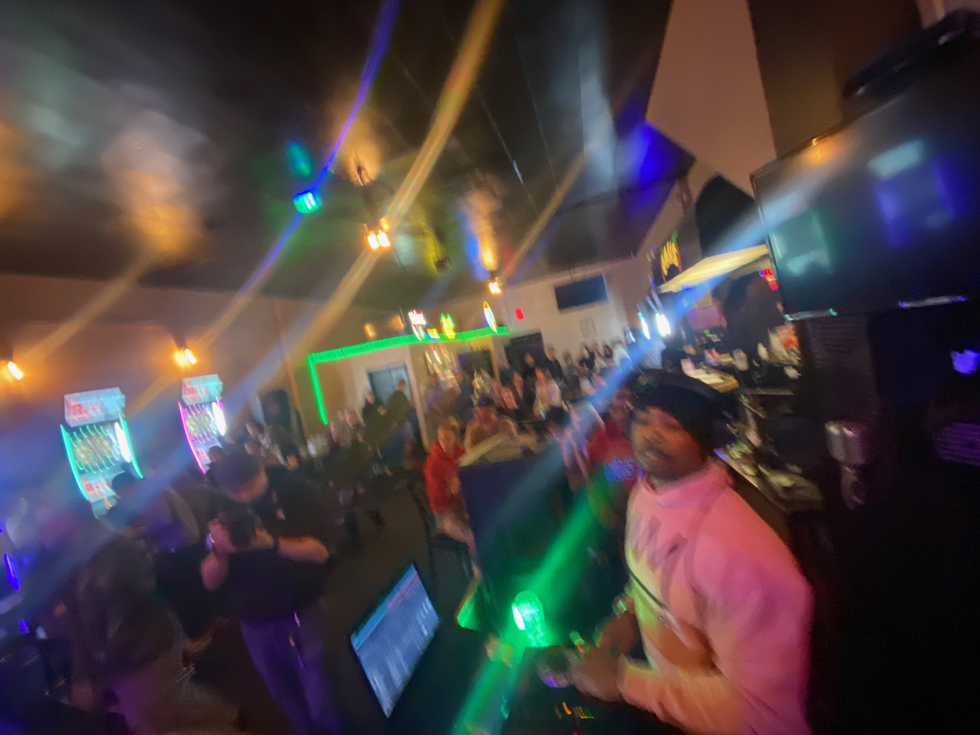 A lively bar scene with people dancing, socializing, and enjoying drinks amid colorful neon lights and a large screen with neon signs.