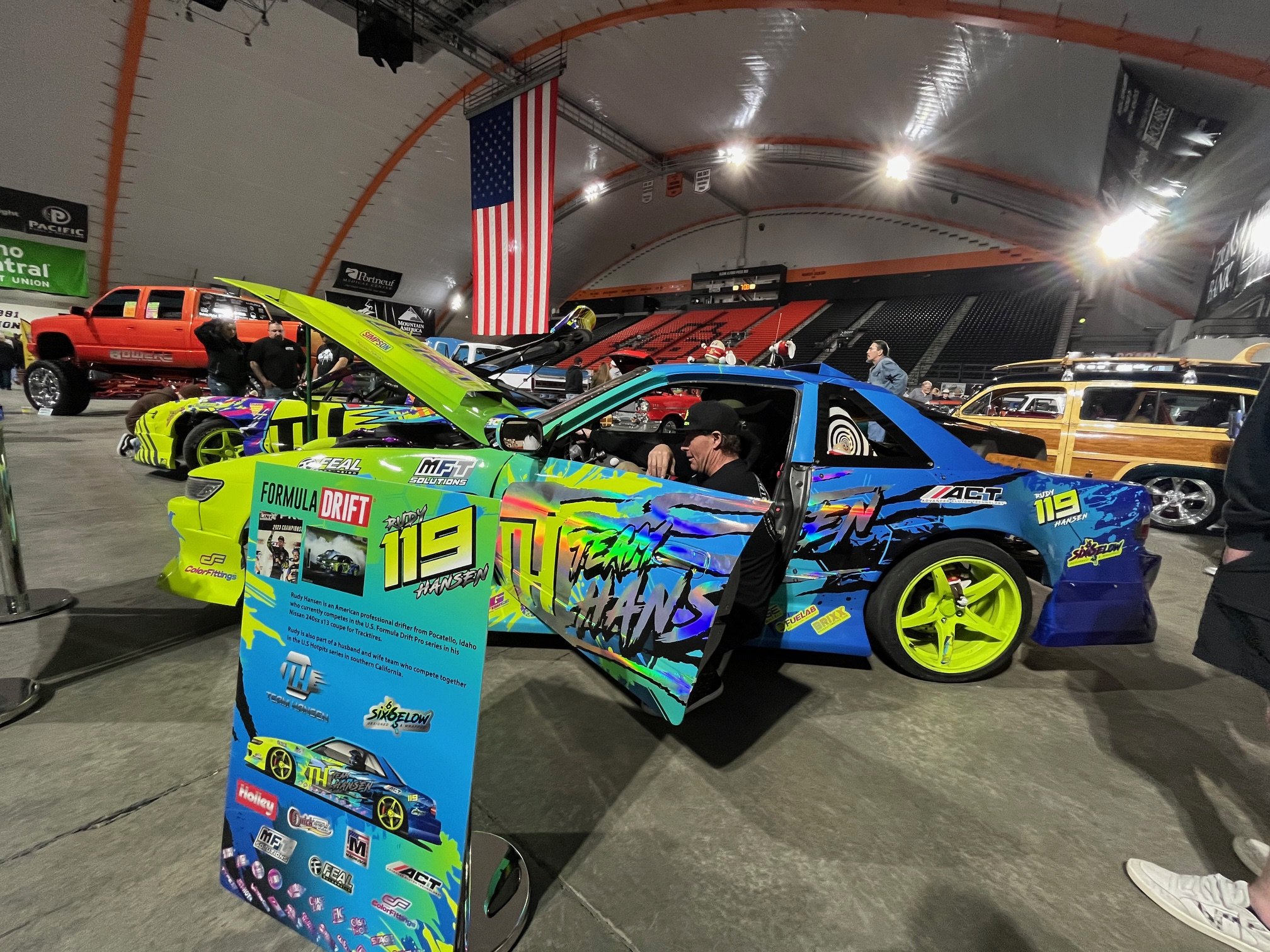 Colorful race car with graphics and the number 119 on the door displayed at an indoor car show, with other vehicles and an American flag in the background.