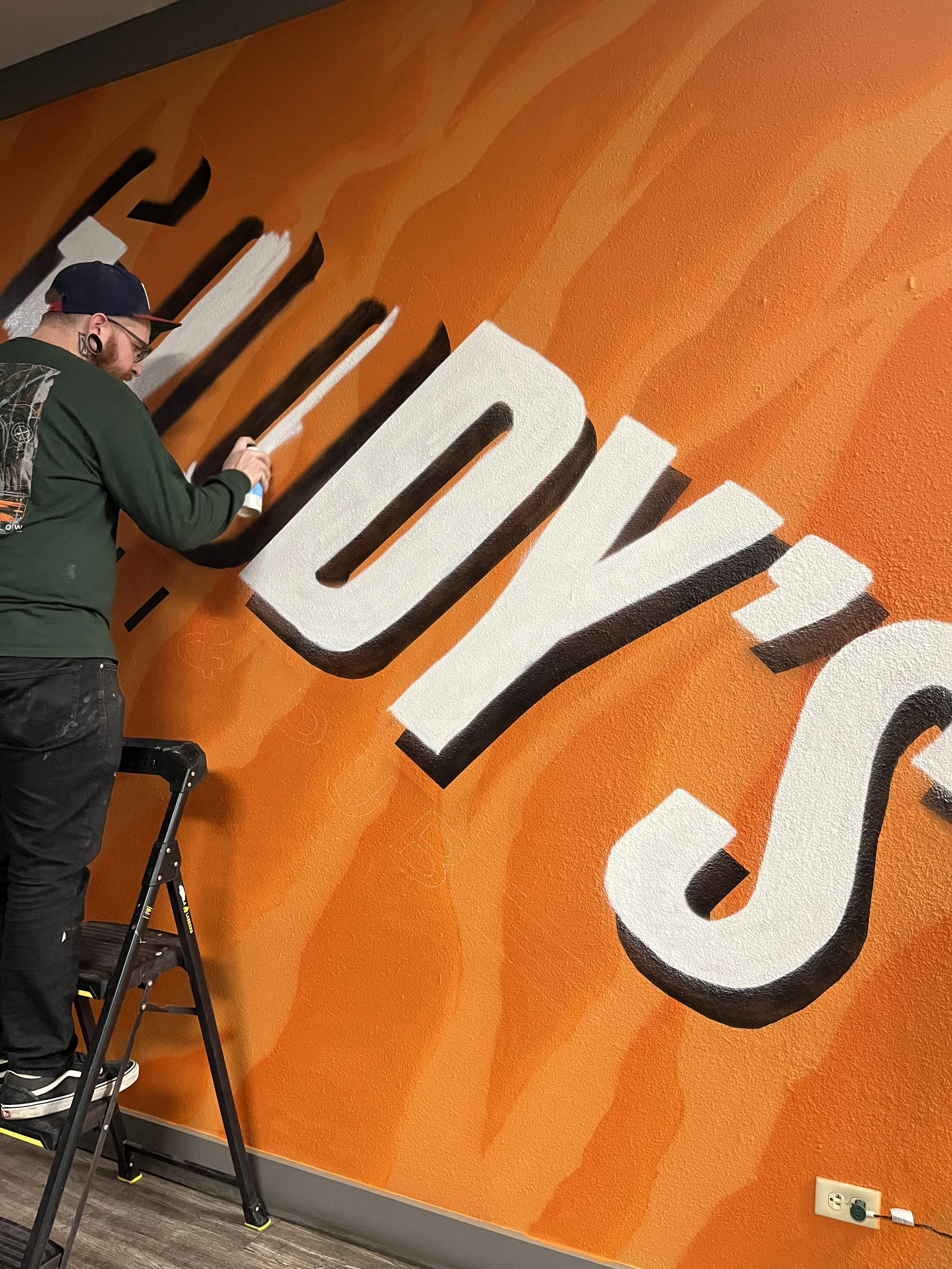 A man is standing on a step ladder painting a large wall mural. The mural is orange with large white and black stylized letters.