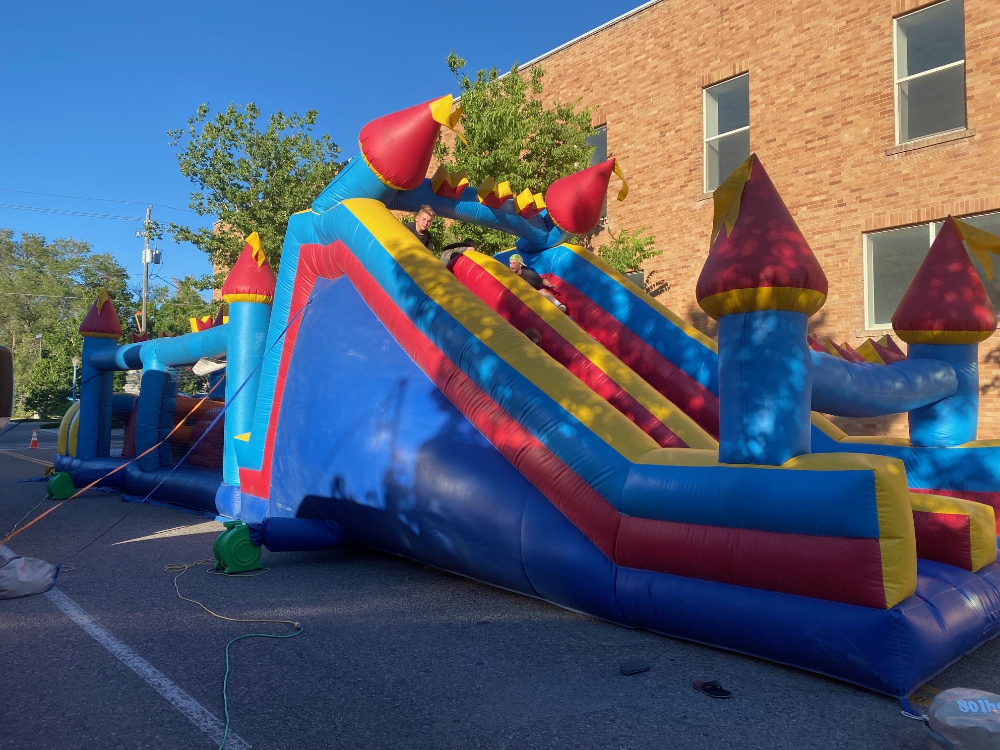 Colorful inflatable bounce house with castle theme, featuring red and yellow turrets and blue walls, set up outdoors on pavement near a brick building and trees on a clear day.