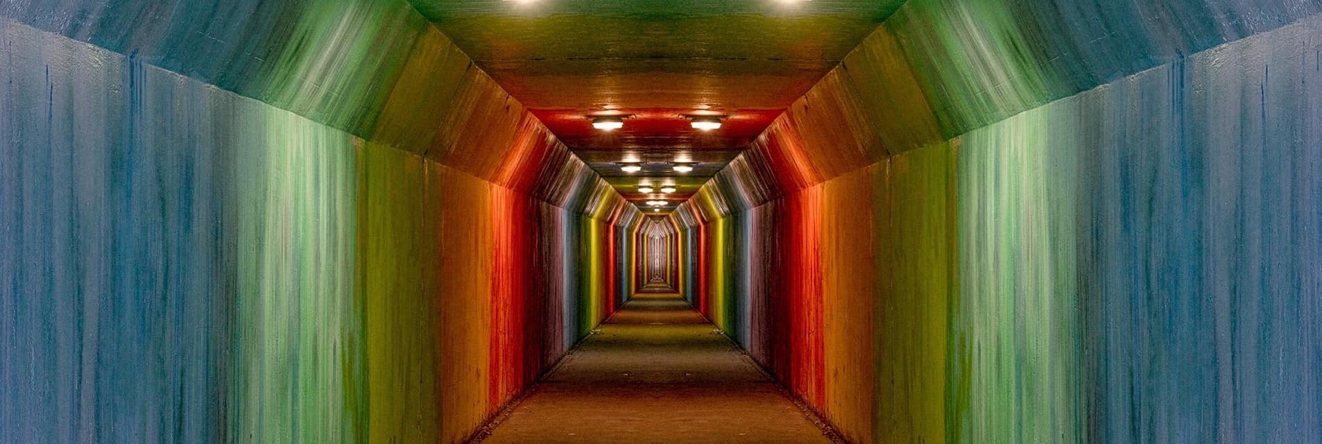 A colorful underground tunnel with walls painted in rainbow colors, illuminated by overhead lights, creating a vibrant and inviting pathway.