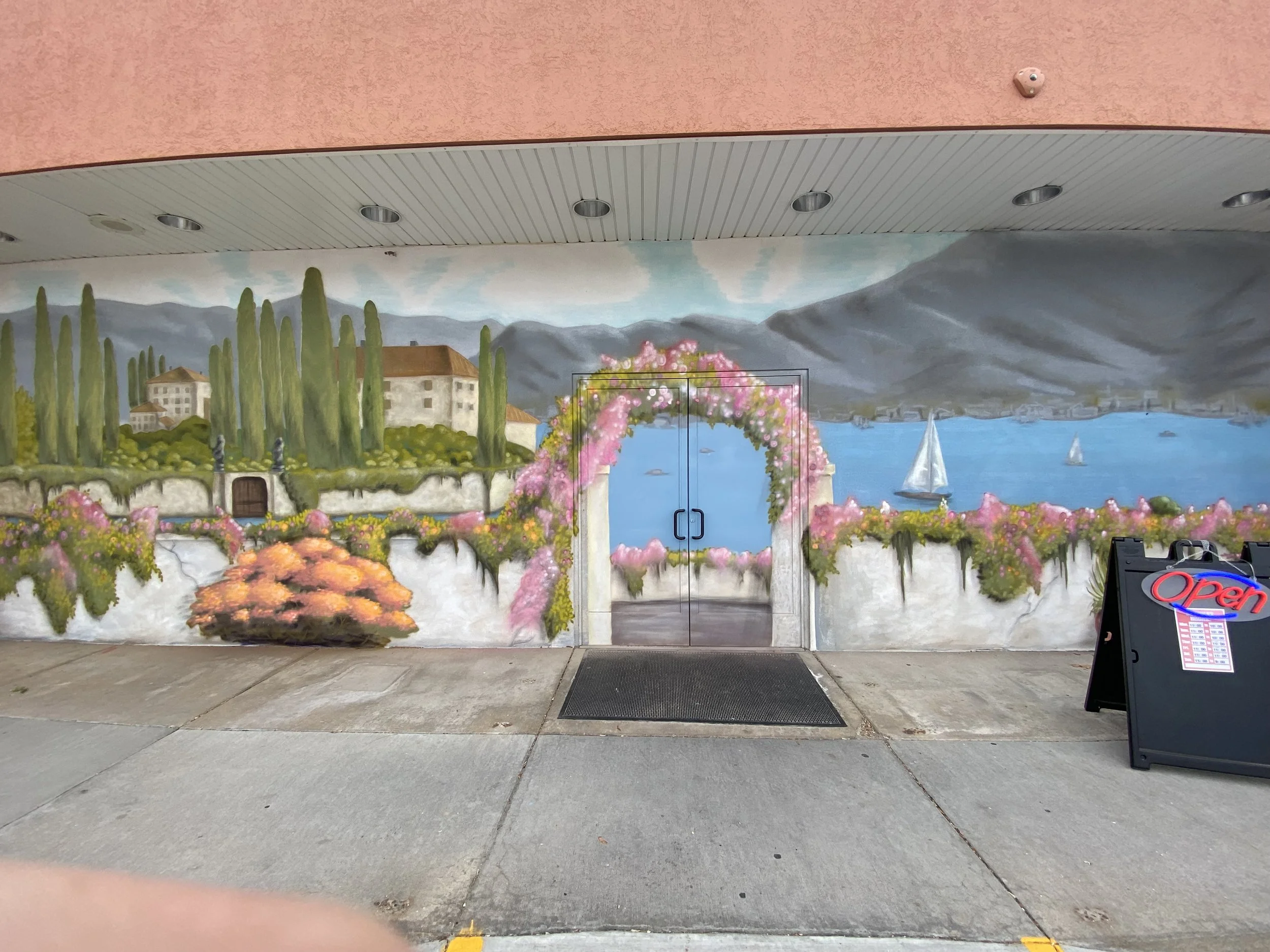 A painted mural on a building exterior showing a scenic landscape with mountains, water, buildings, trees, pink flowering plants, and sailboats, with a glass double door in the center.