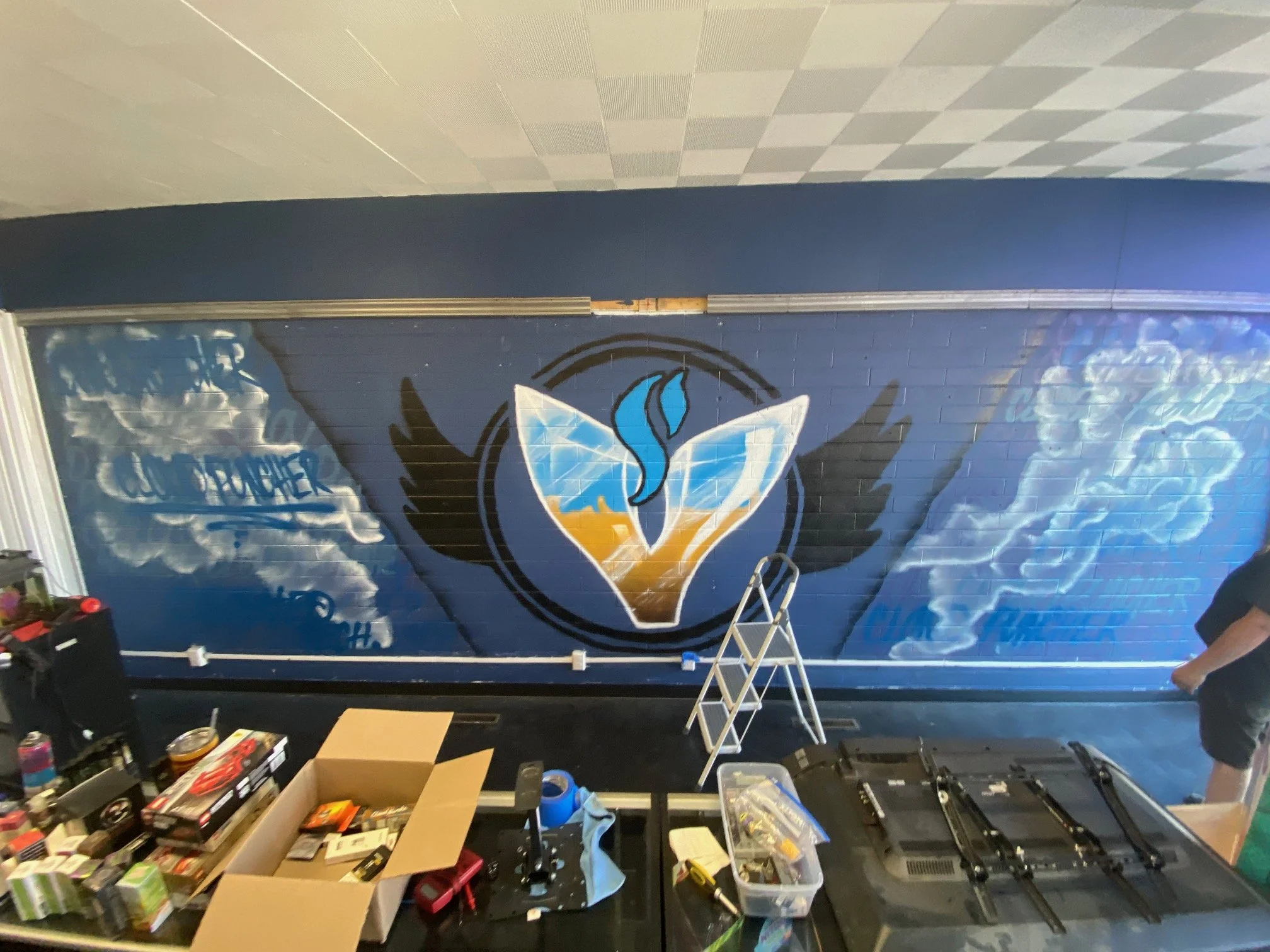 A room with a large wall mural of a logo, some boxes and items on a table, and various tools and equipment on the floor, indicating a workspace or workshop setup.