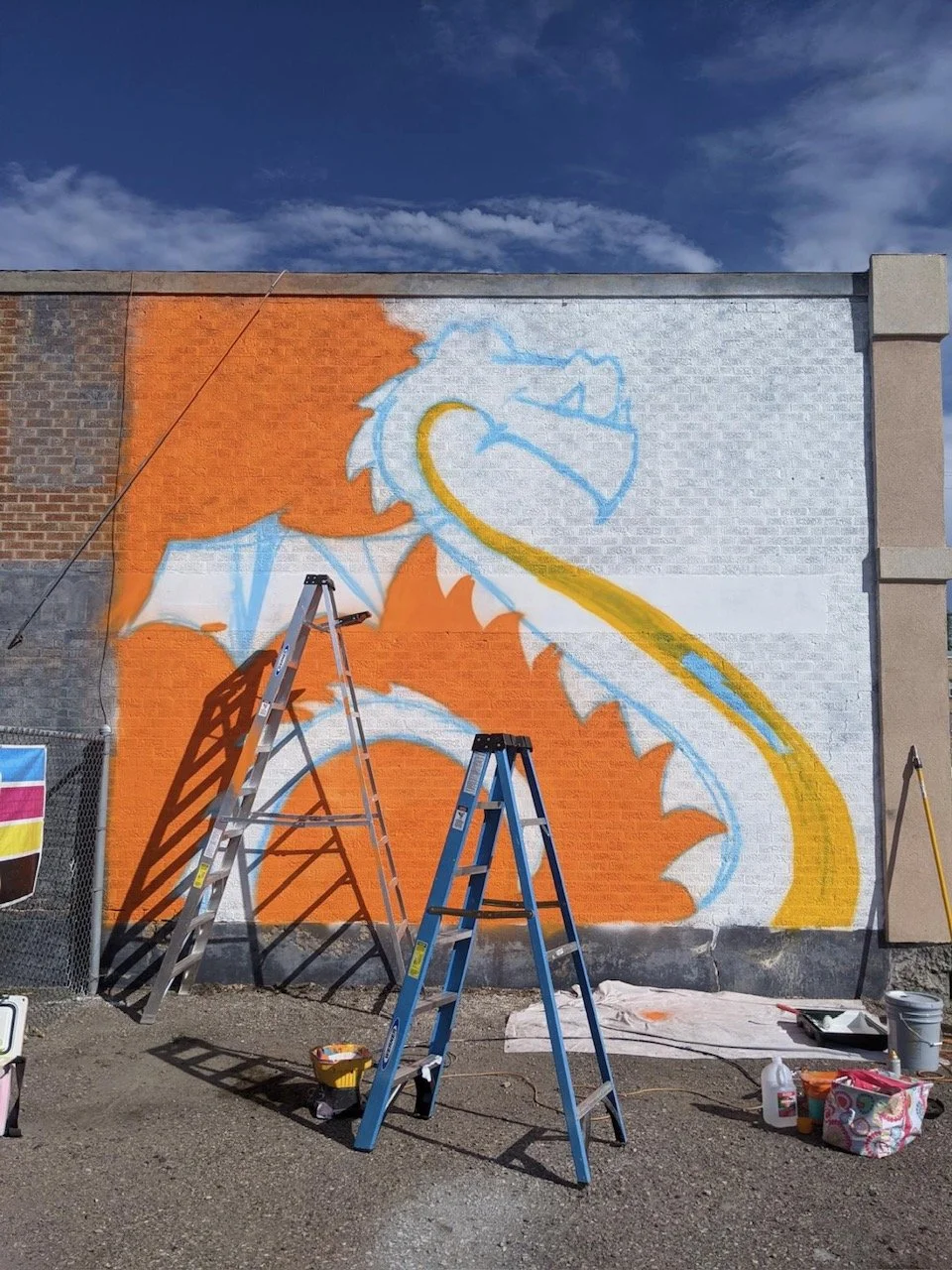 A mural of a cartoon dragon in progress on a brick wall, with ladders and painting supplies in the foreground, under a partly cloudy sky.